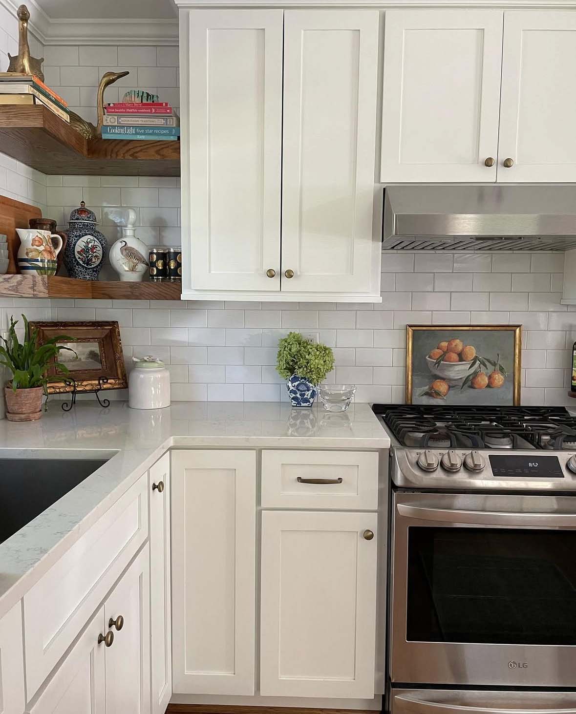 Modern white kitchen corner with stainless steel stove, white cabinets, wooden shelves with decorative items and cookbooks, and a small plant on the countertop.