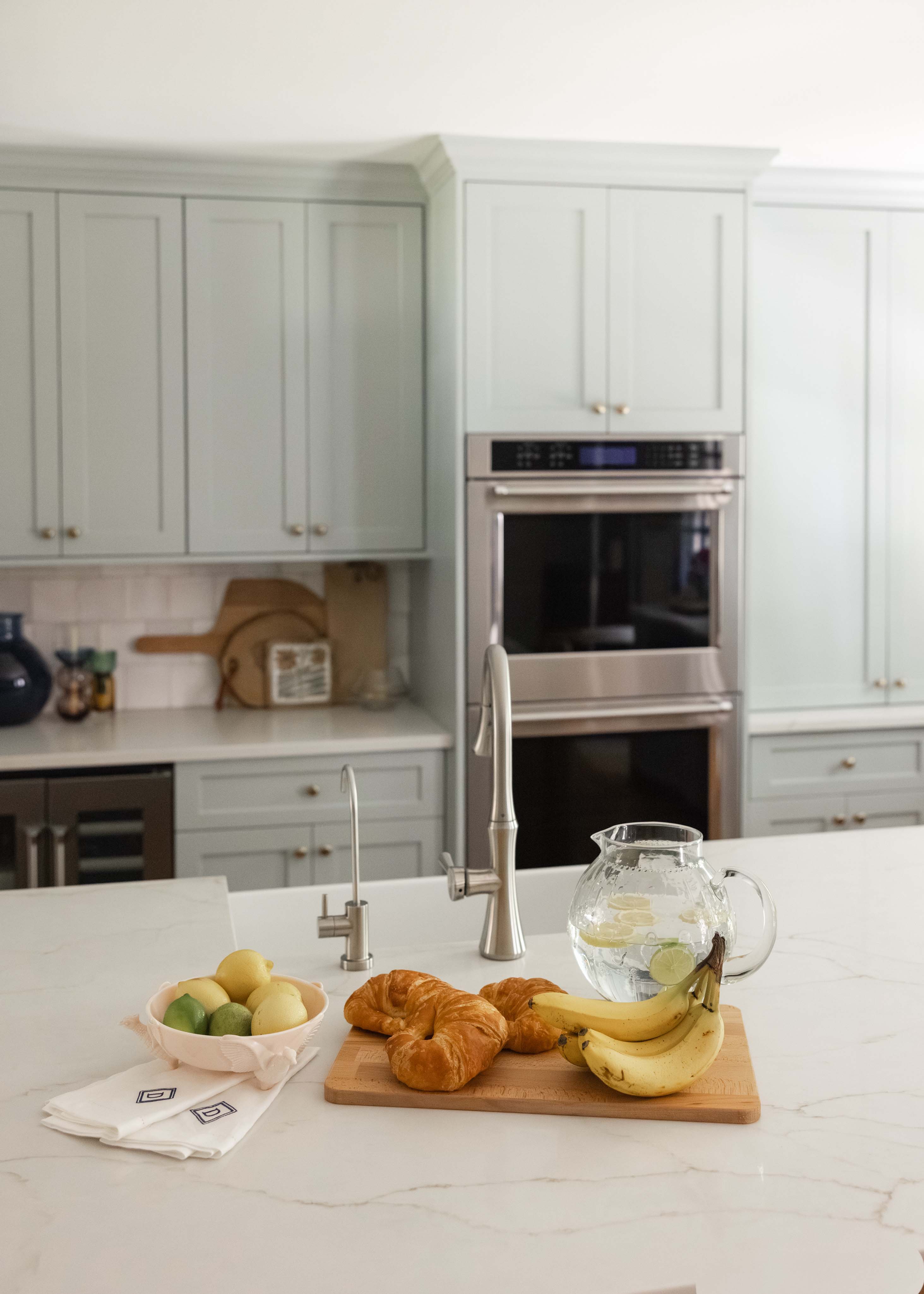 Modern kitchen island with a wooden cutting board holding croissants and bananas, a glass pitcher with lemon water, and a bowl of lemons and limes on a marble countertop.