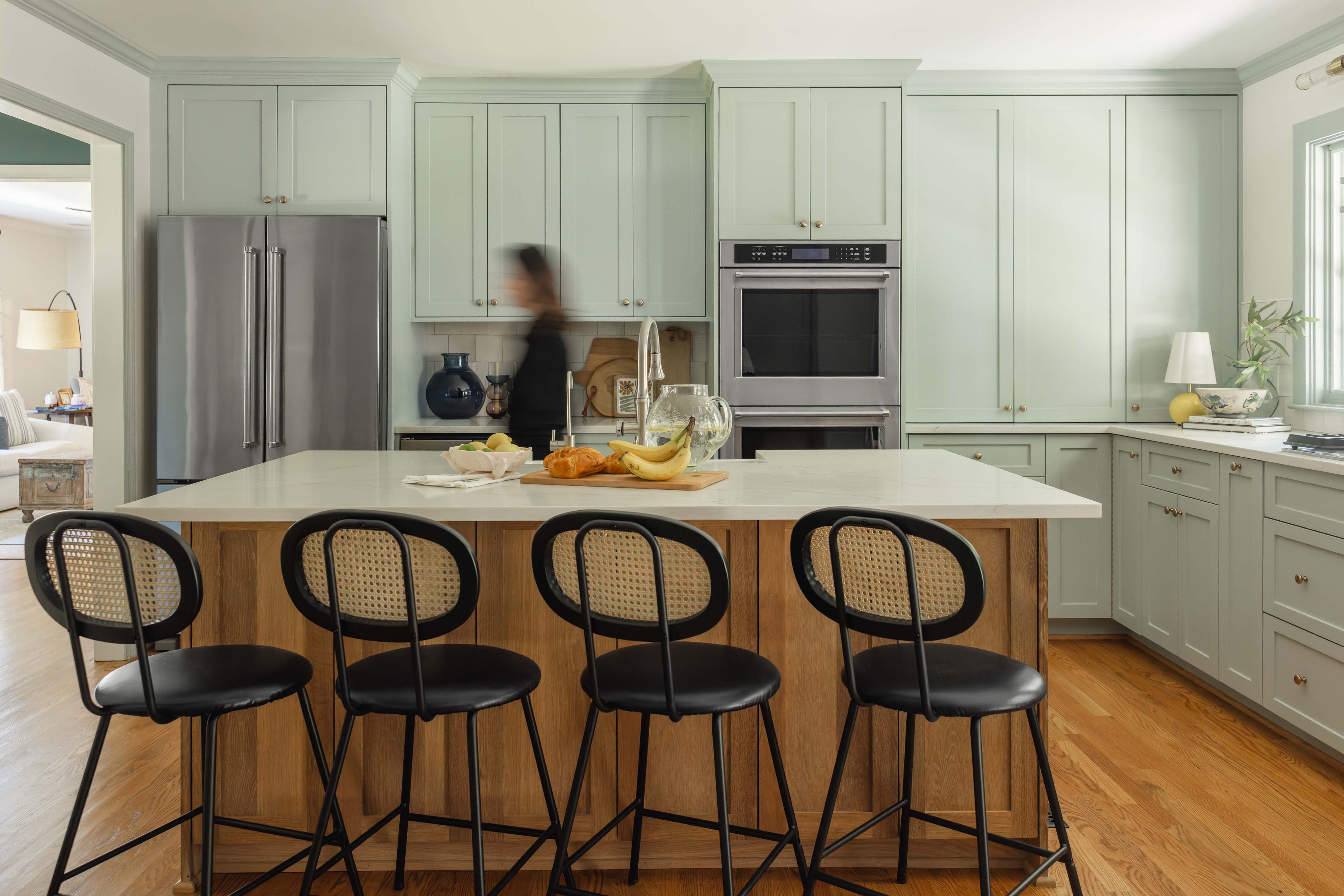 Modern kitchen with light green cabinets, a large island with wooden base and white countertop, four black chairs, and a woman walking in the background.