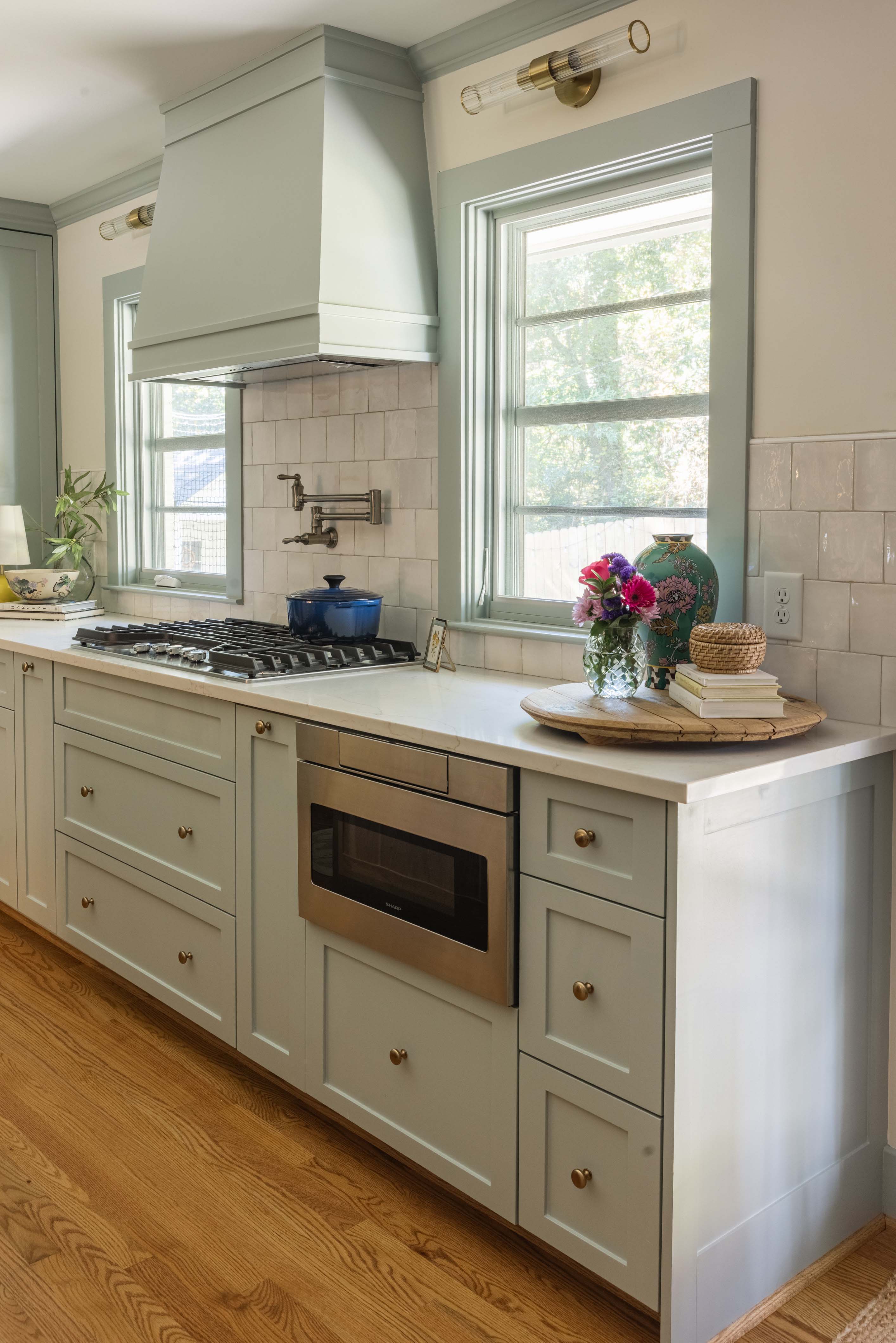 Light green kitchen cabinetry with a stainless steel microwave, gas stove, white countertop, wooden floor, and decorative tray with flowers and a vase by the window.