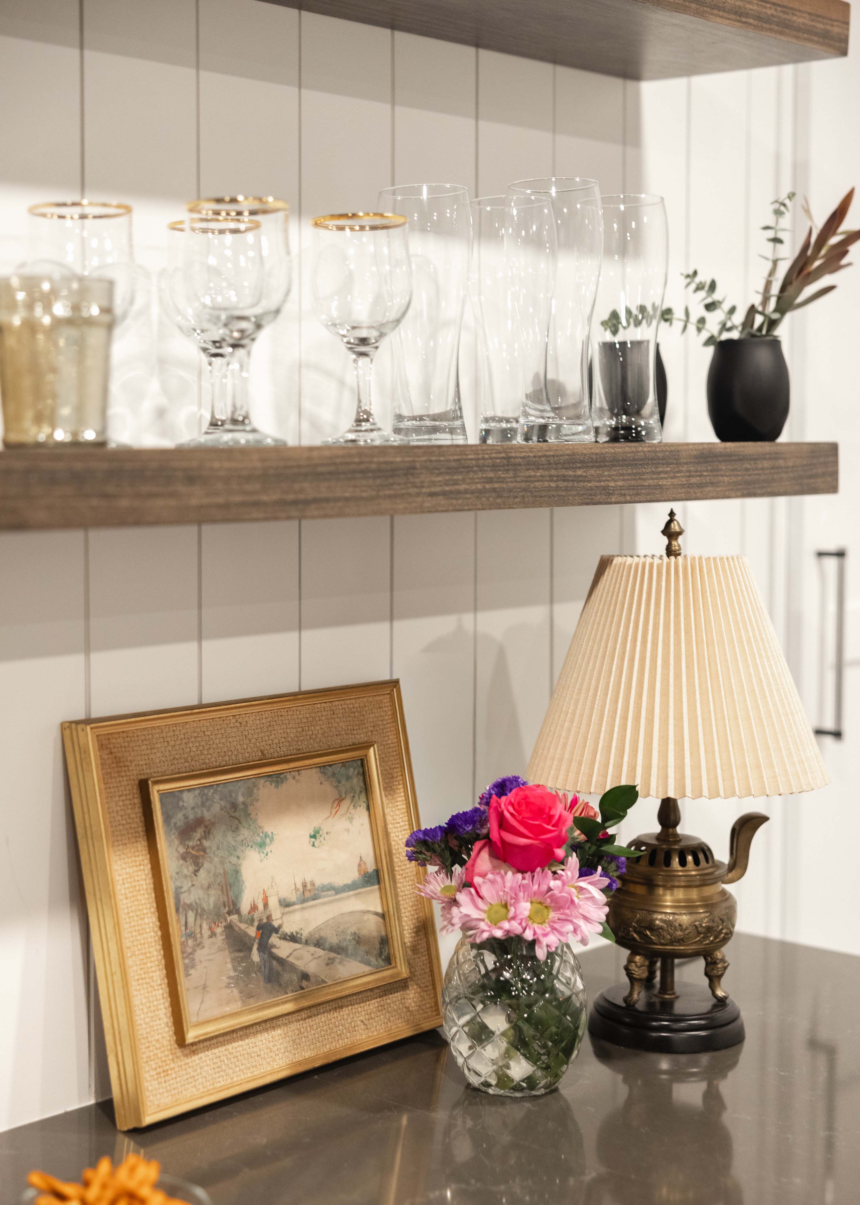Wooden shelf with glassware and black vase above a countertop holding a framed painting, a glass vase with mixed flowers, and a vintage brass lamp with pleated shade.