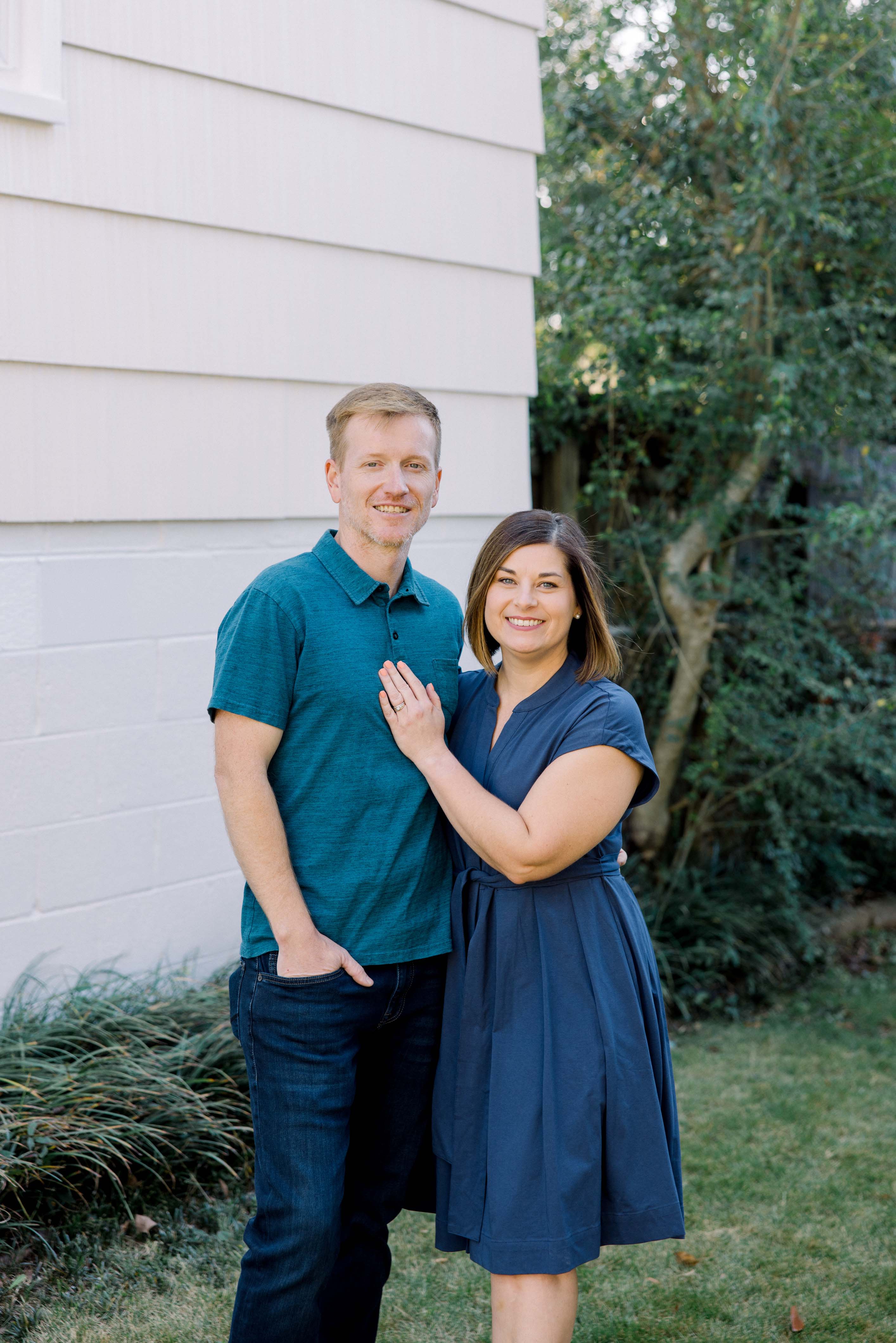 Smiling couple standing outdoors with woman wearing a navy dress and man in a teal polo and jeans.