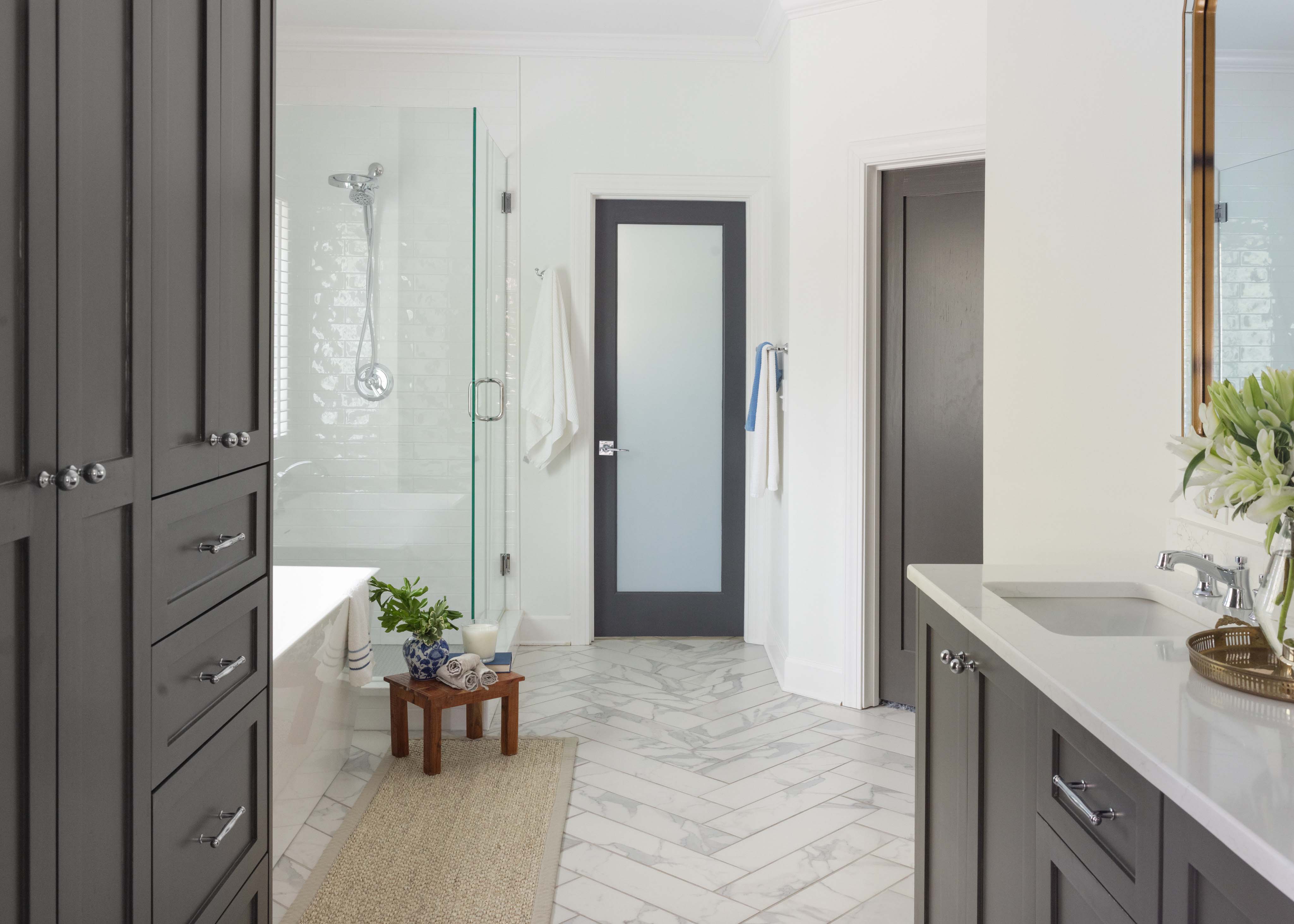 Bright modern bathroom with gray cabinets, glass shower, white bathtub, and marble tile floor, featuring a small wooden stool with rolled towels and a potted plant.