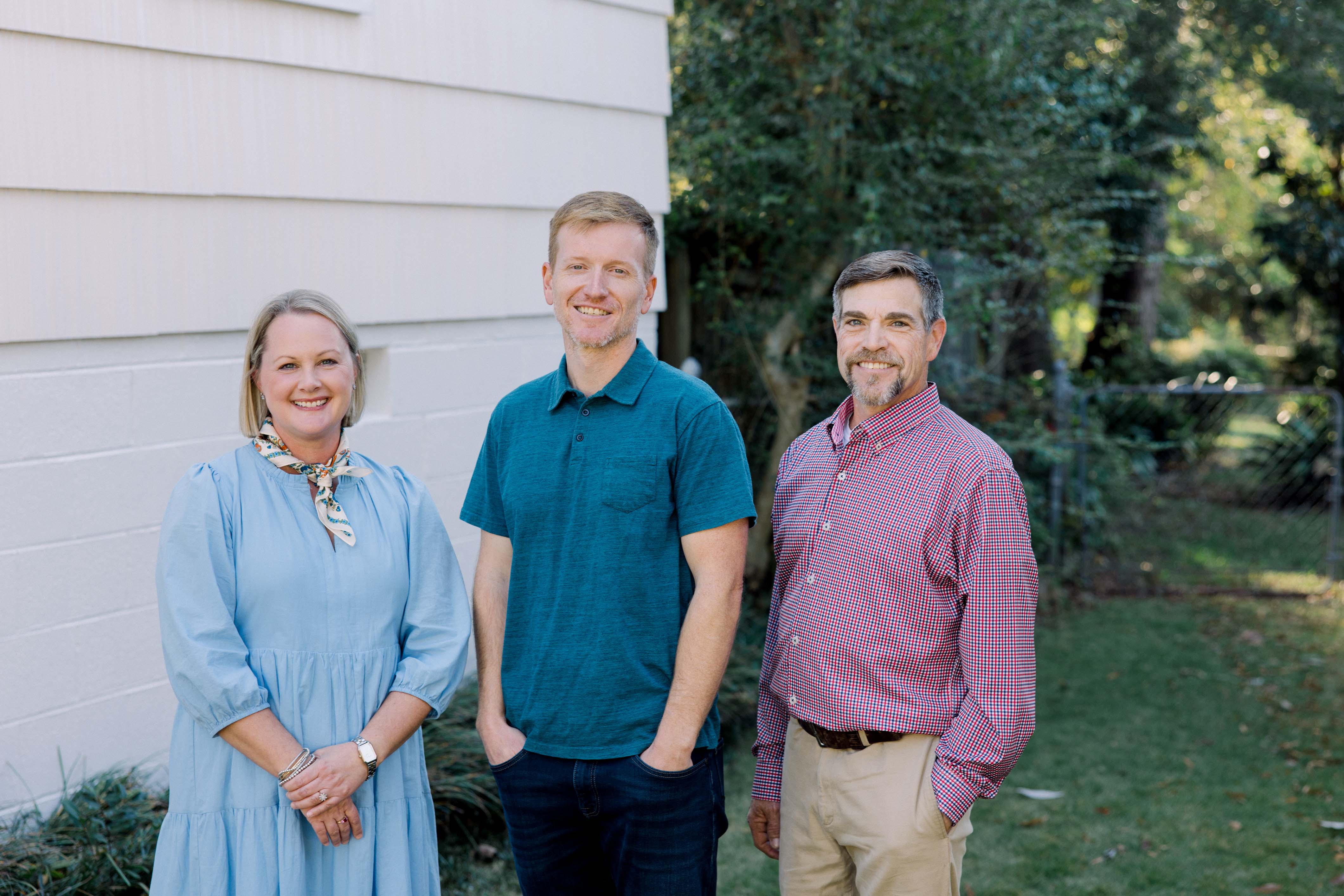 Three adults smiling outdoors in front of a house, standing on grass with trees and a fence in the background.