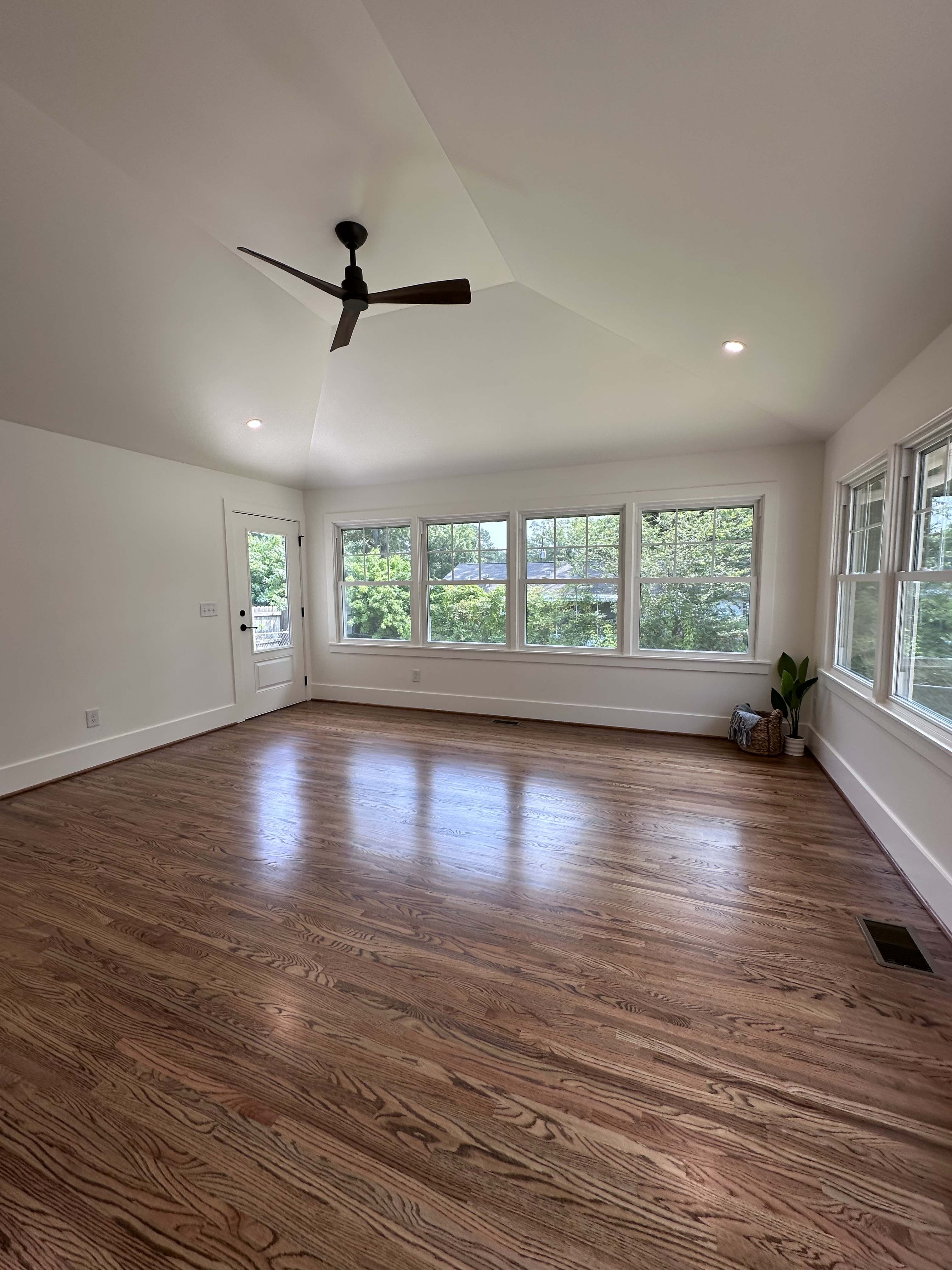 Bright empty sunroom with hardwood floors, white walls, large windows showing greenery outside, ceiling fan, and a plant with a basket in the corner.