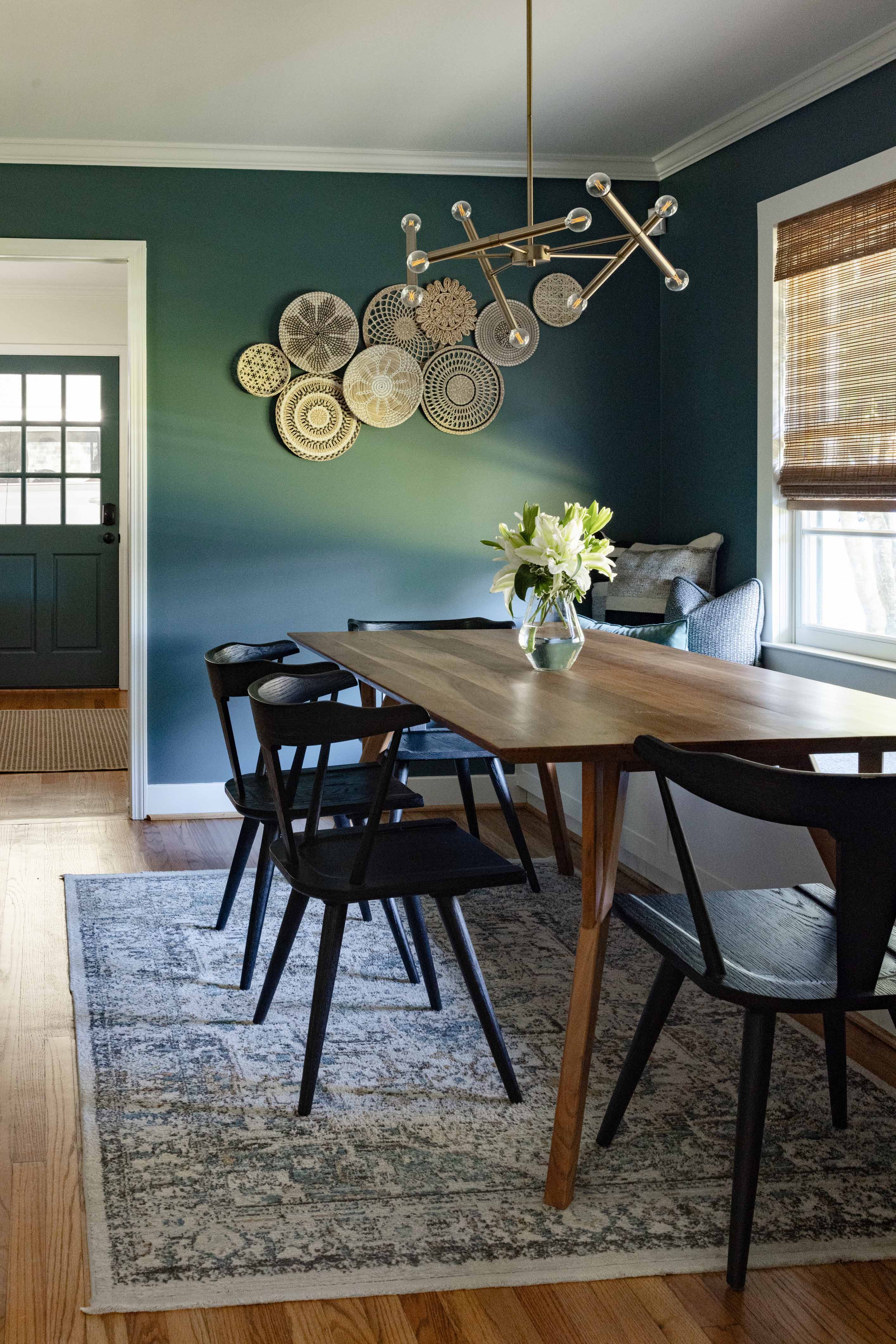 Dining room with wooden table, black chairs, green walls with decorative woven wall hangings, and a modern brass chandelier.