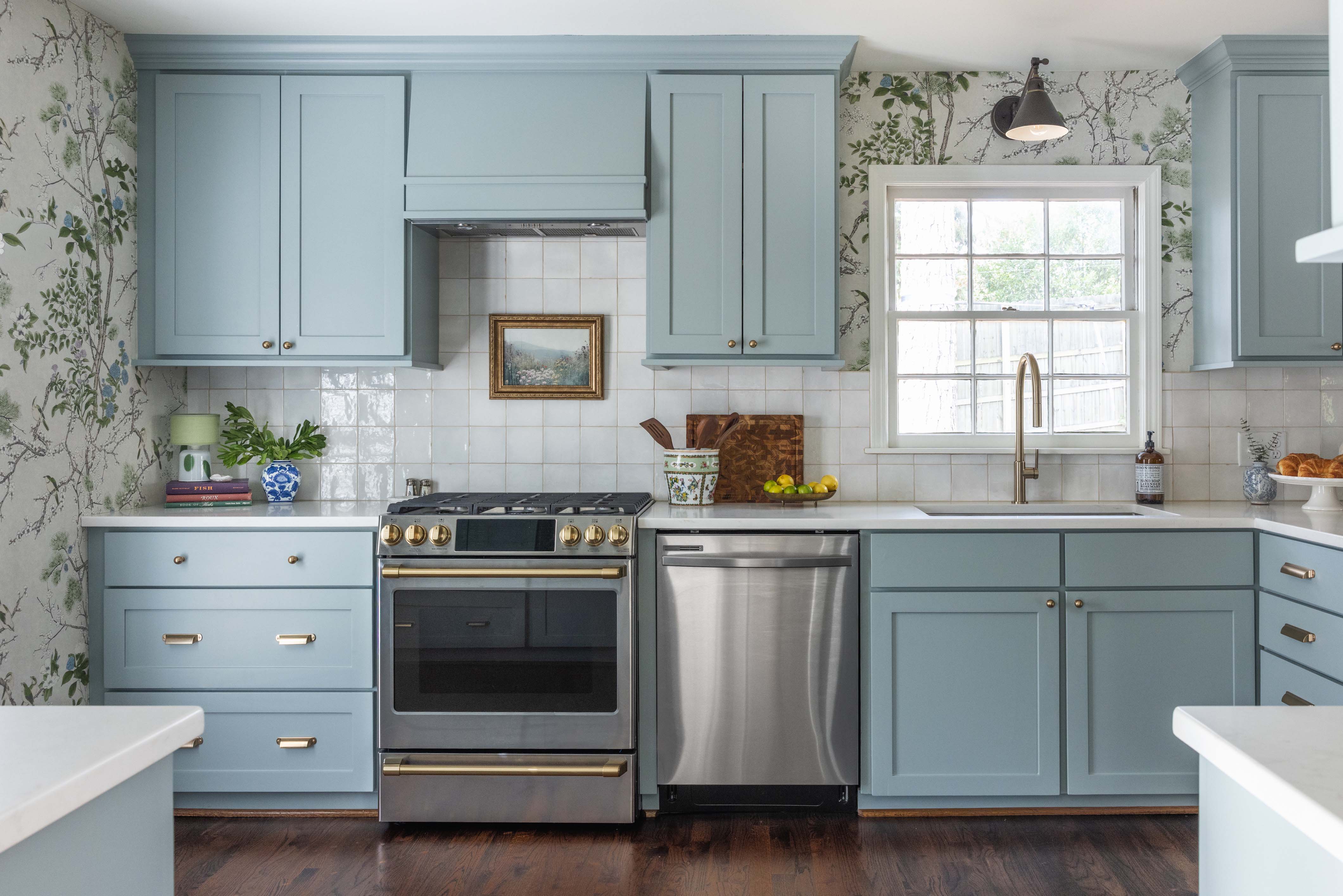 Bright kitchen with light blue cabinets, stainless steel stove and dishwasher, floral wallpaper, white countertops, and a window over the sink.