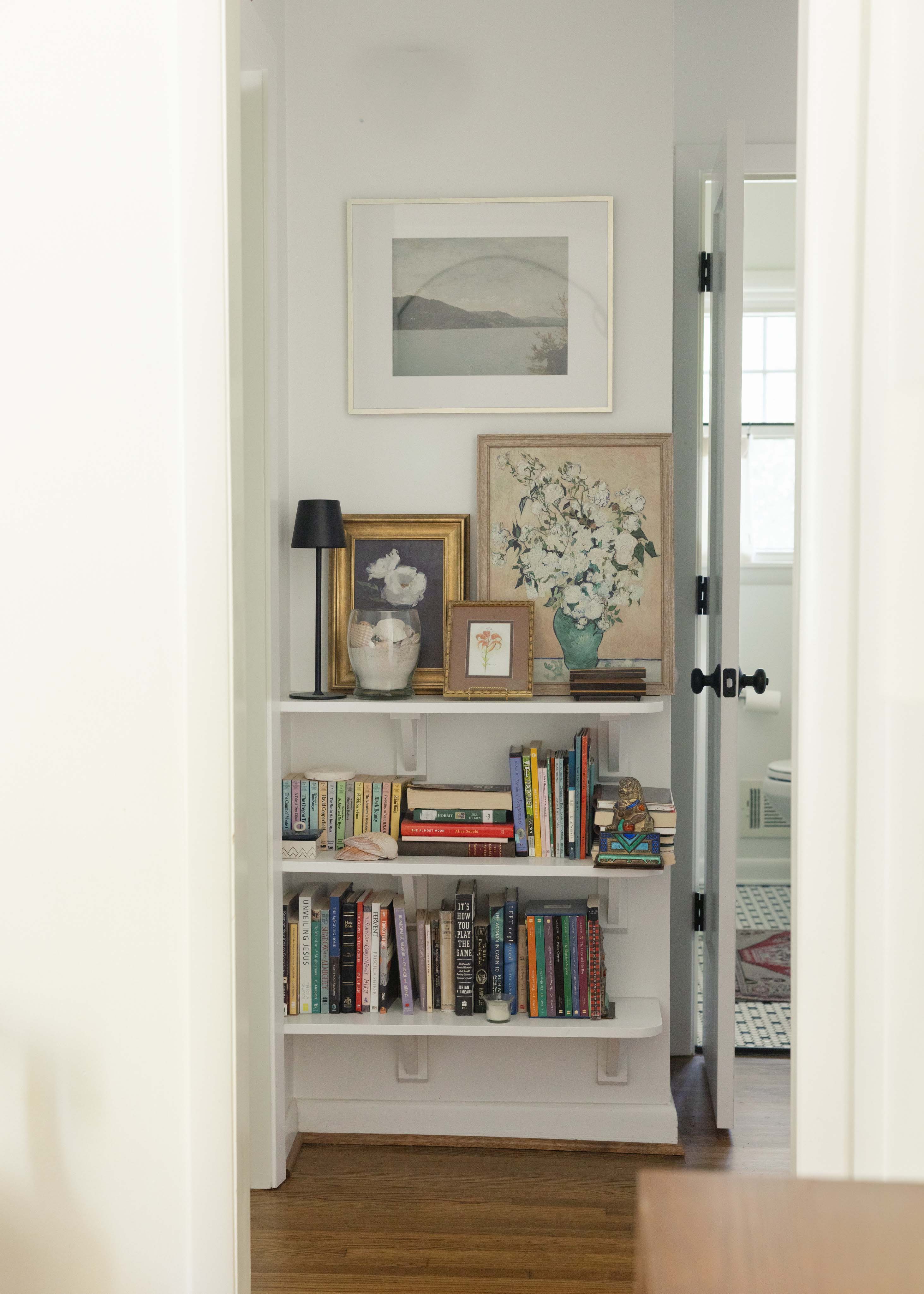 White built-in bookshelf filled with books, framed floral artwork, a table lamp, and decorative items, located in a hallway with a partially open door to a bathroom.