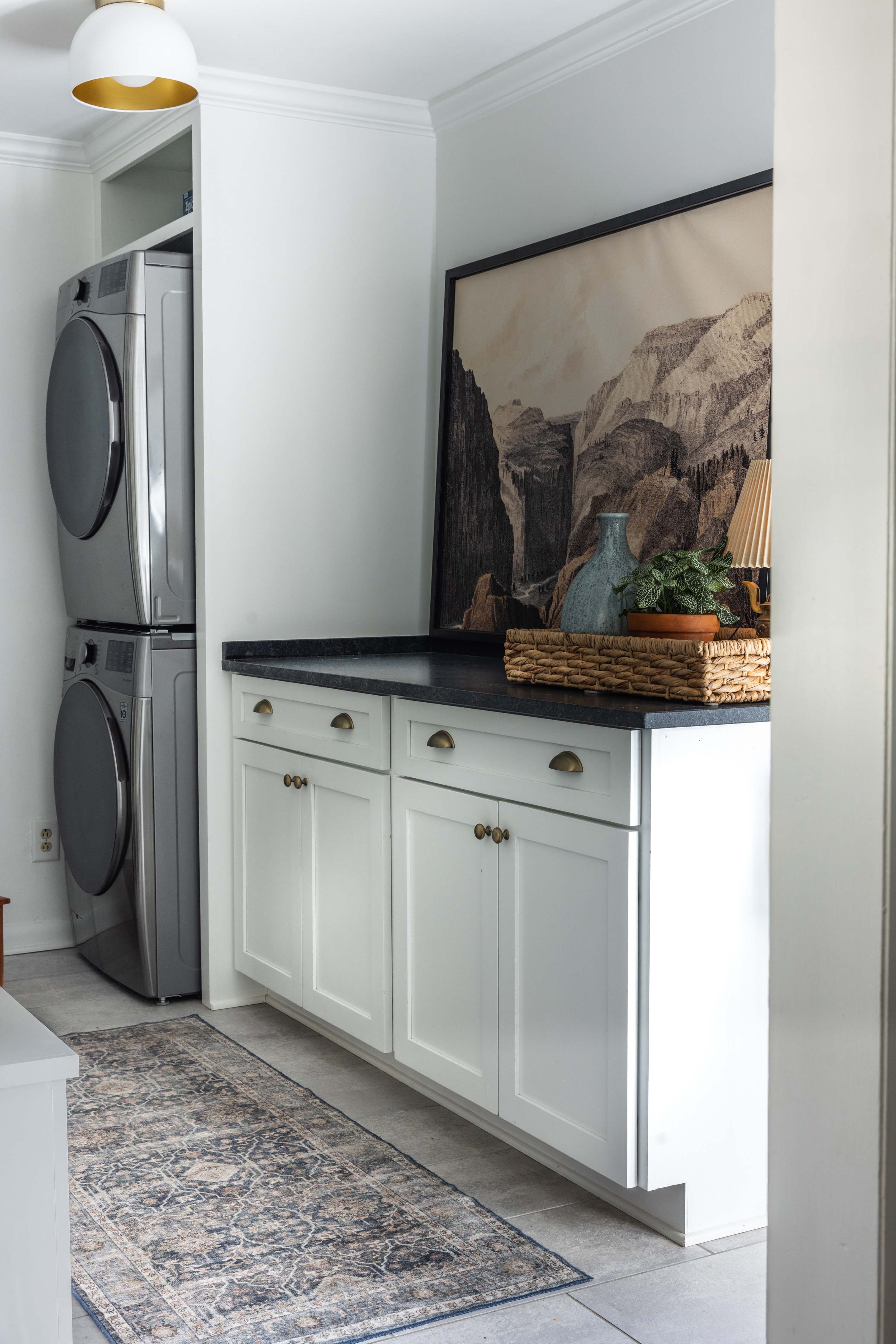 Laundry room with stacked silver washer and dryer, white cabinets with black countertop, and a decorative tray with plant, vase, and lamp.