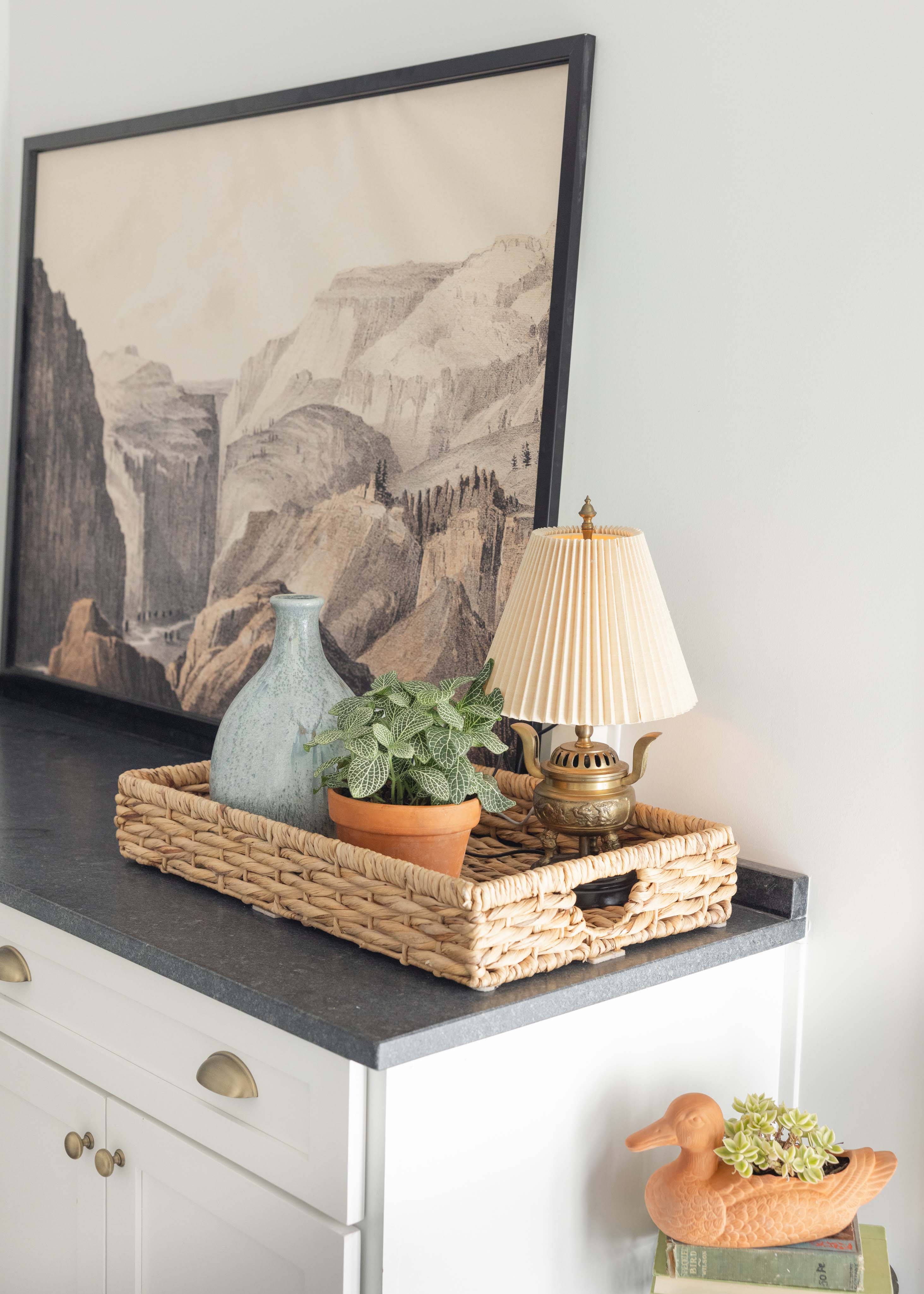 Wicker tray on dark countertop holding a green ceramic vase, a small potted plant, and a brass lamp with a pleated shade, with a framed mountain landscape artwork in the background and a duck-shaped planter on books below.