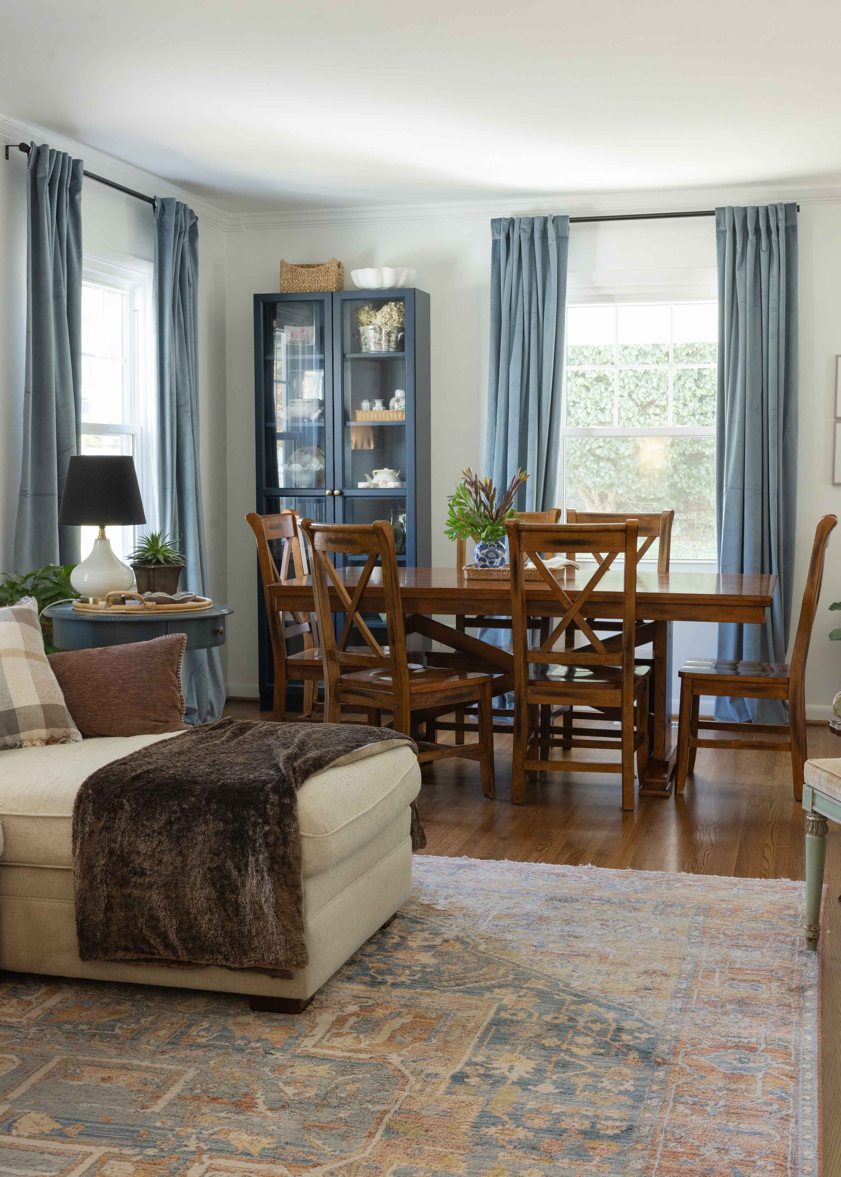 Wooden dining table with six matching chairs, a small blue and white vase with greenery on the table, blue curtains framing a window, and a cabinet with dishes on the left.
