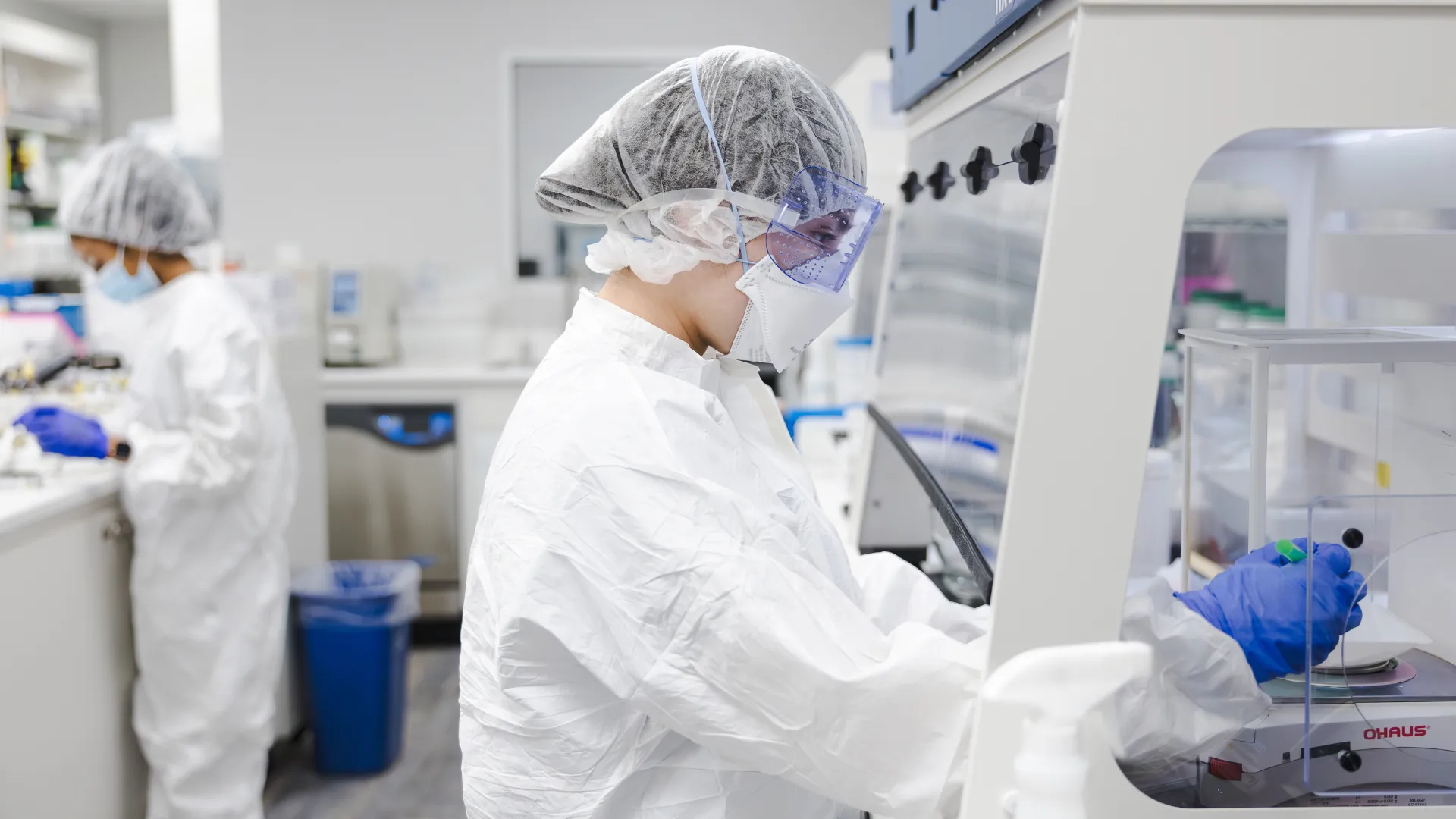 Two lab technicians in white protective suits working in a sterile laboratory setting.