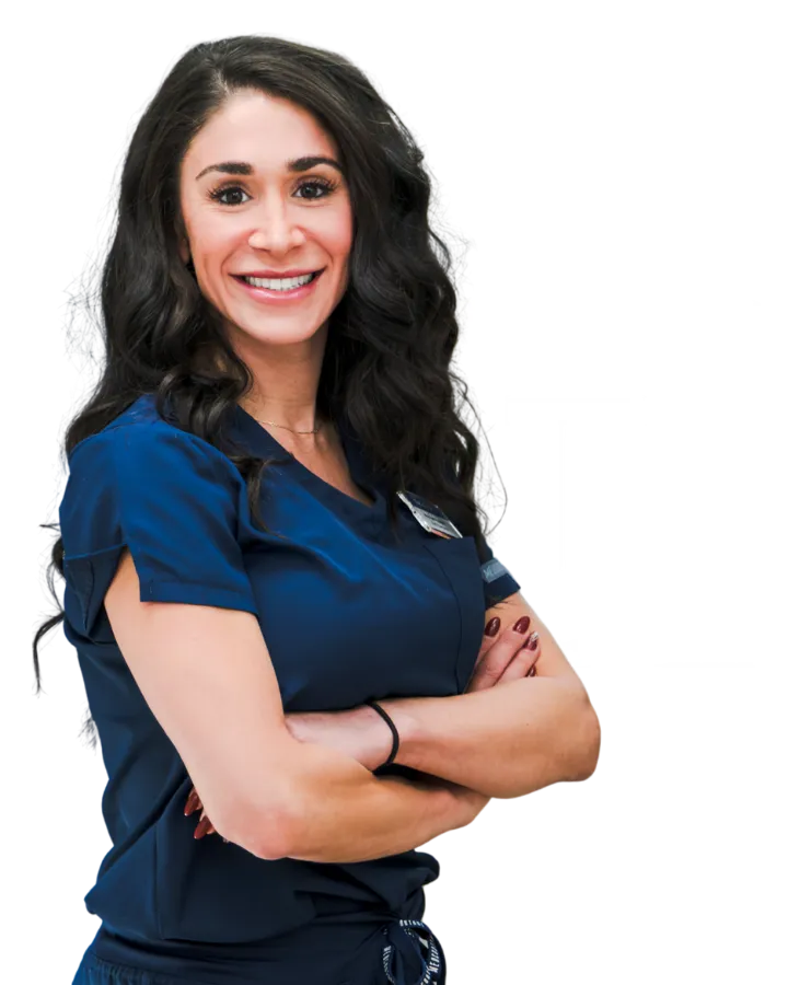 Smiling female medical worker with long dark curly hair wearing navy blue scrubs and arms crossed.