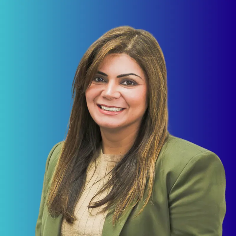 Smiling woman with long brown hair wearing a beige top and olive green blazer on blue gradient background.