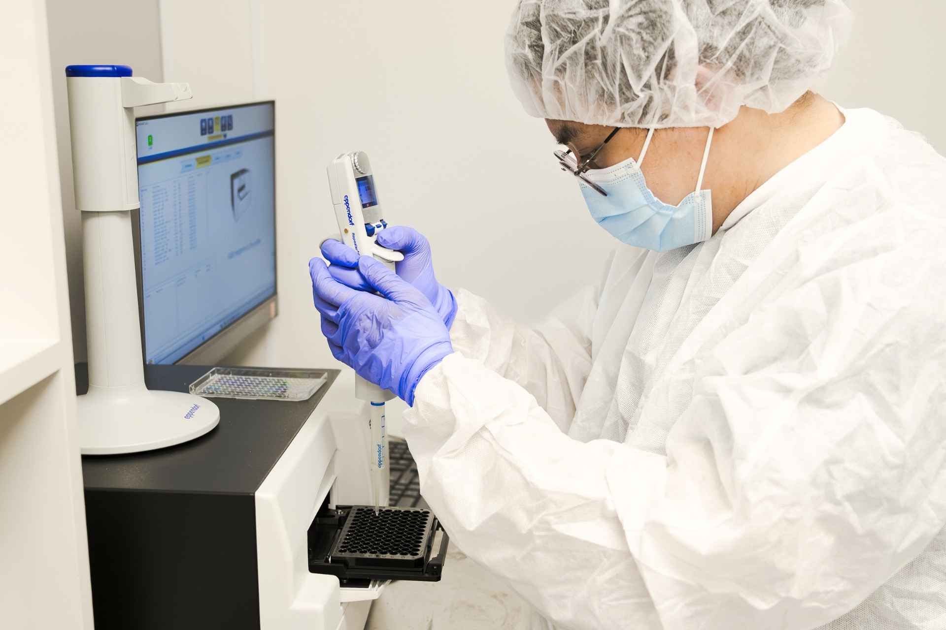 Scientist in protective gear using a micropipette to transfer liquid into a multi-well plate in a laboratory.