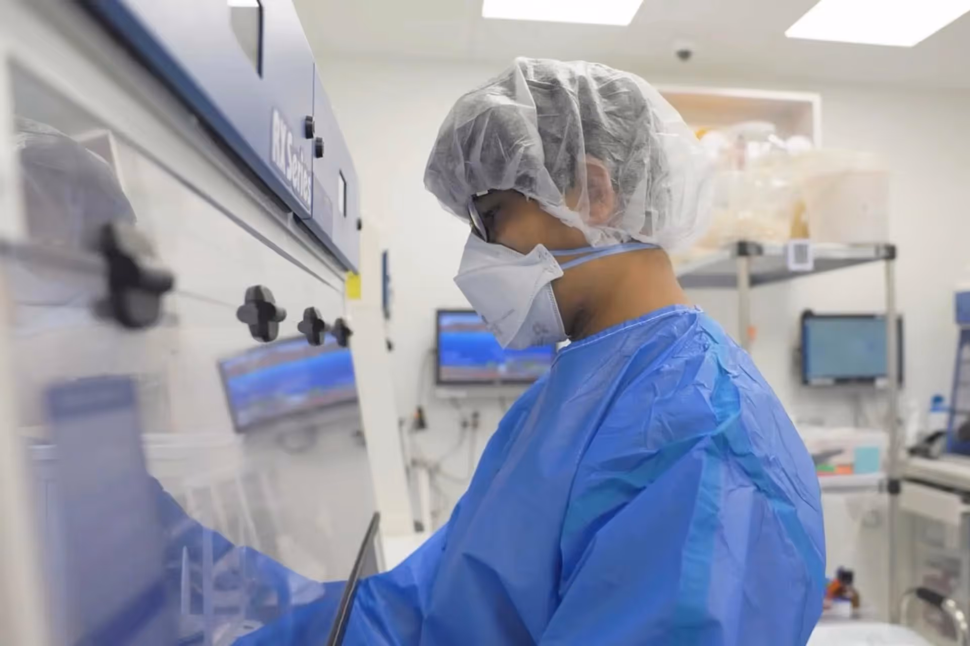 Pharmacist in protective gown, hairnet, and mask working with a laboratory hood in a pharmacy lab.
