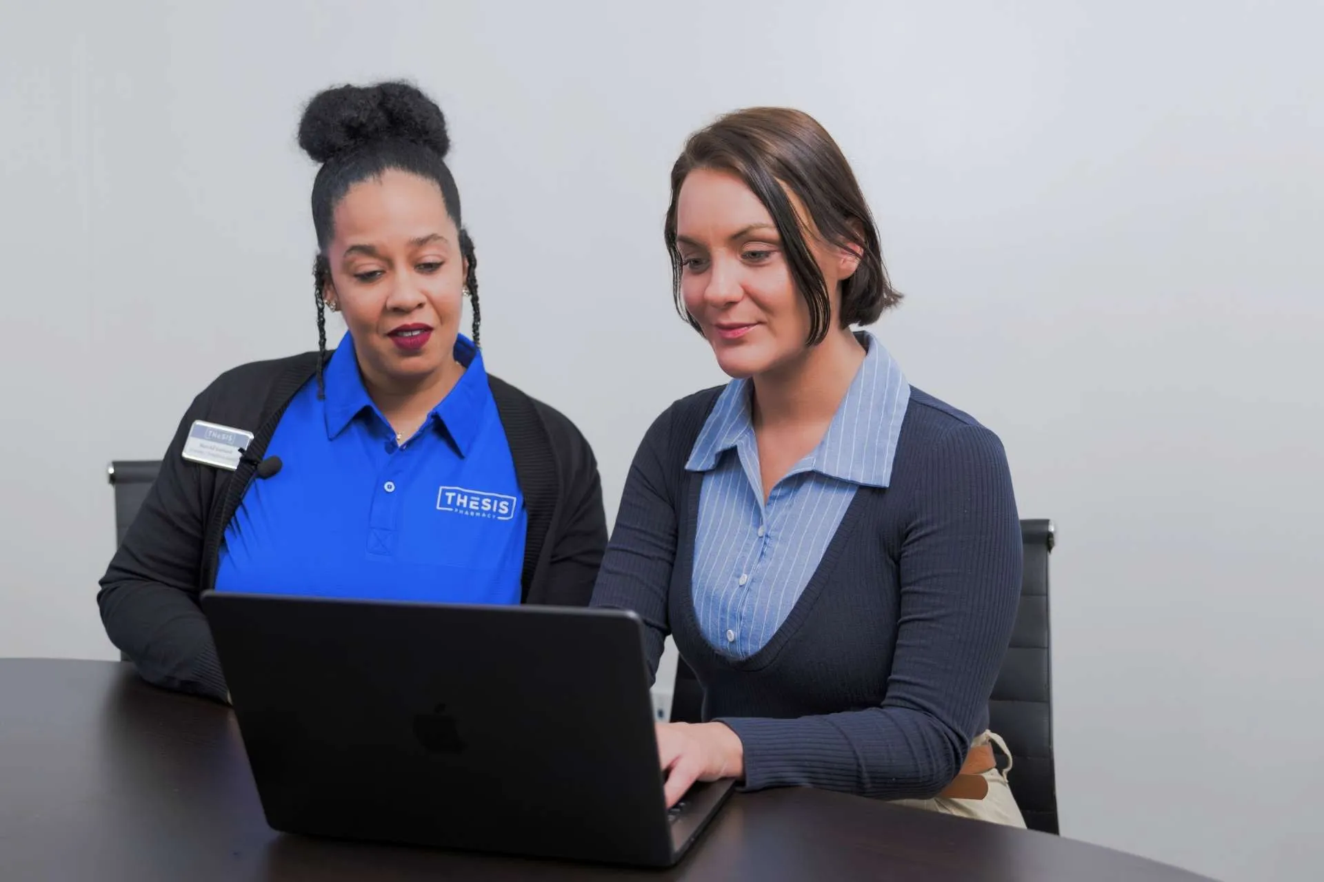 Two women sitting at a table, looking at a laptop, one wearing a blue Thesis Pharmacy shirt.