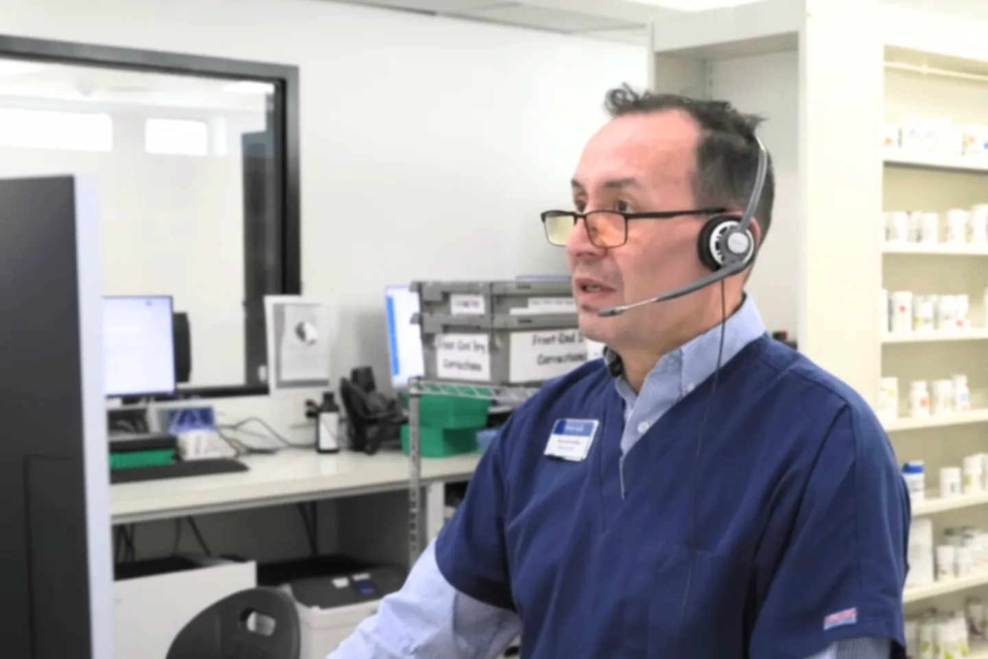 A pharmacist wearing glasses and headset working in a pharmacy with medicine bottles on shelves.