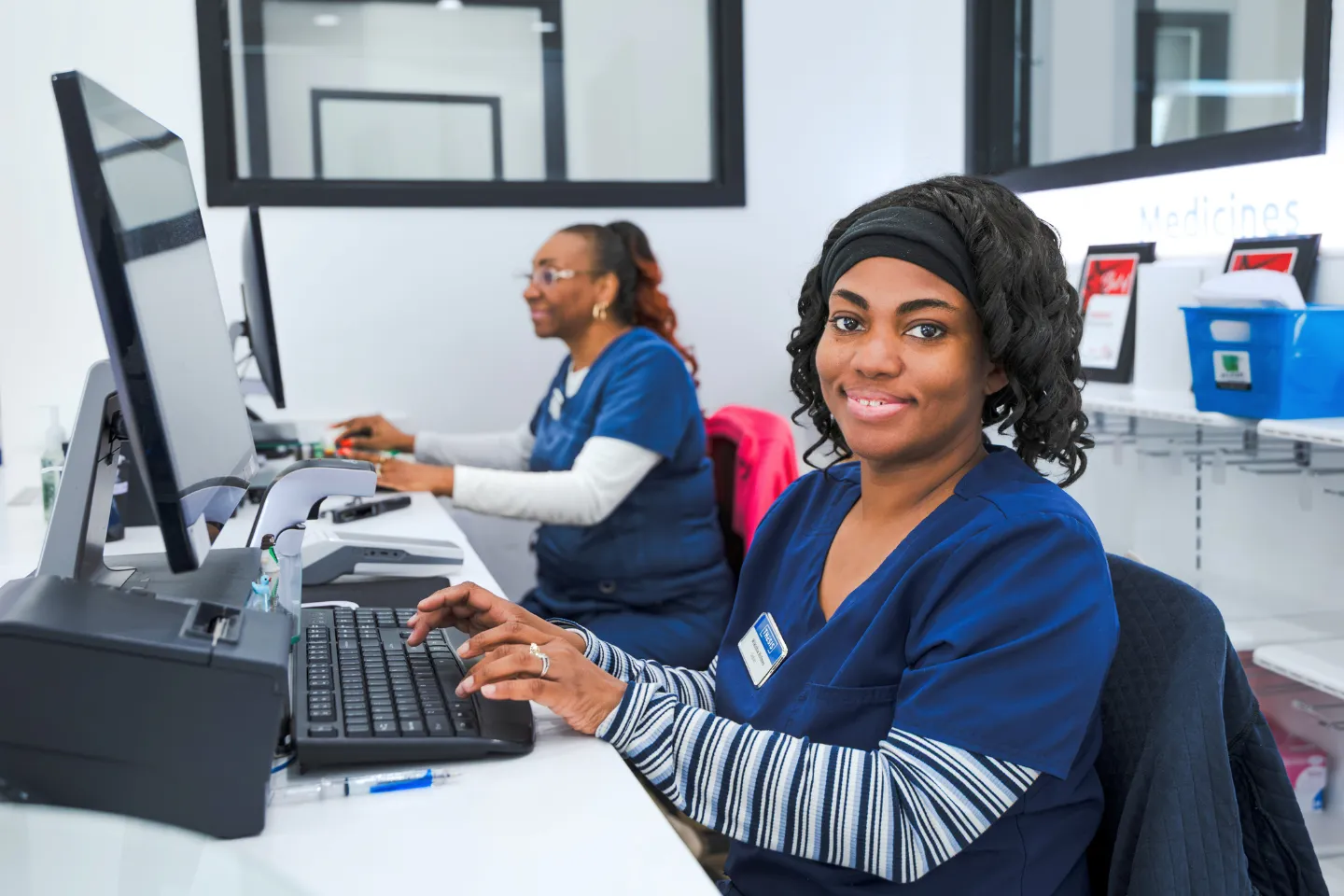 Two healthcare workers in blue scrubs at a pharmacy computer, one smiling at the camera.