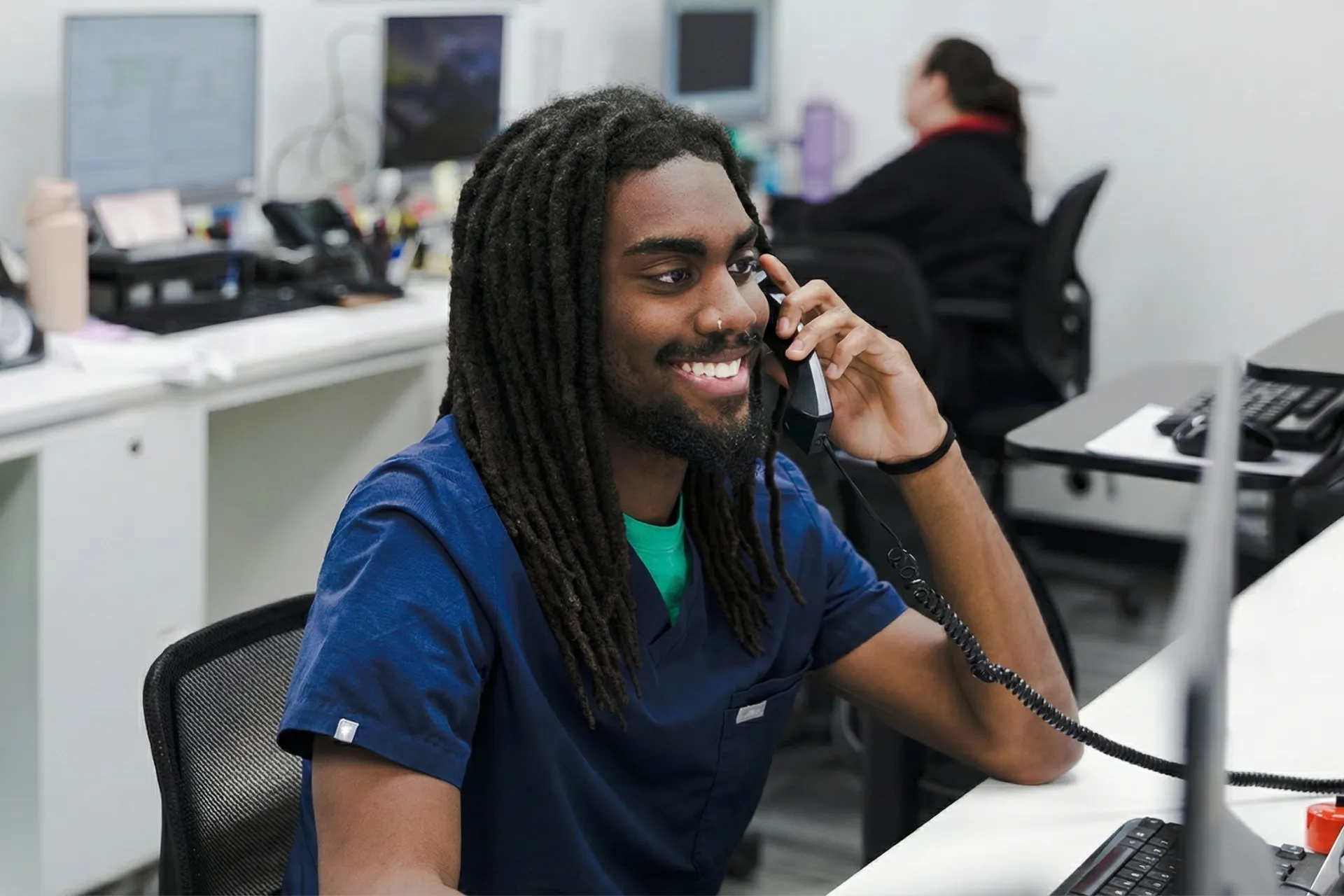 Smiling man in blue scrubs talking on a phone at a desk in an office with computer monitors.