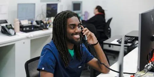 Smiling man with dreadlocks wearing scrubs talks on phone at a computer in an office.