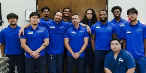 Group of eleven men in matching blue work shirts posing indoors, smiling and standing close together.