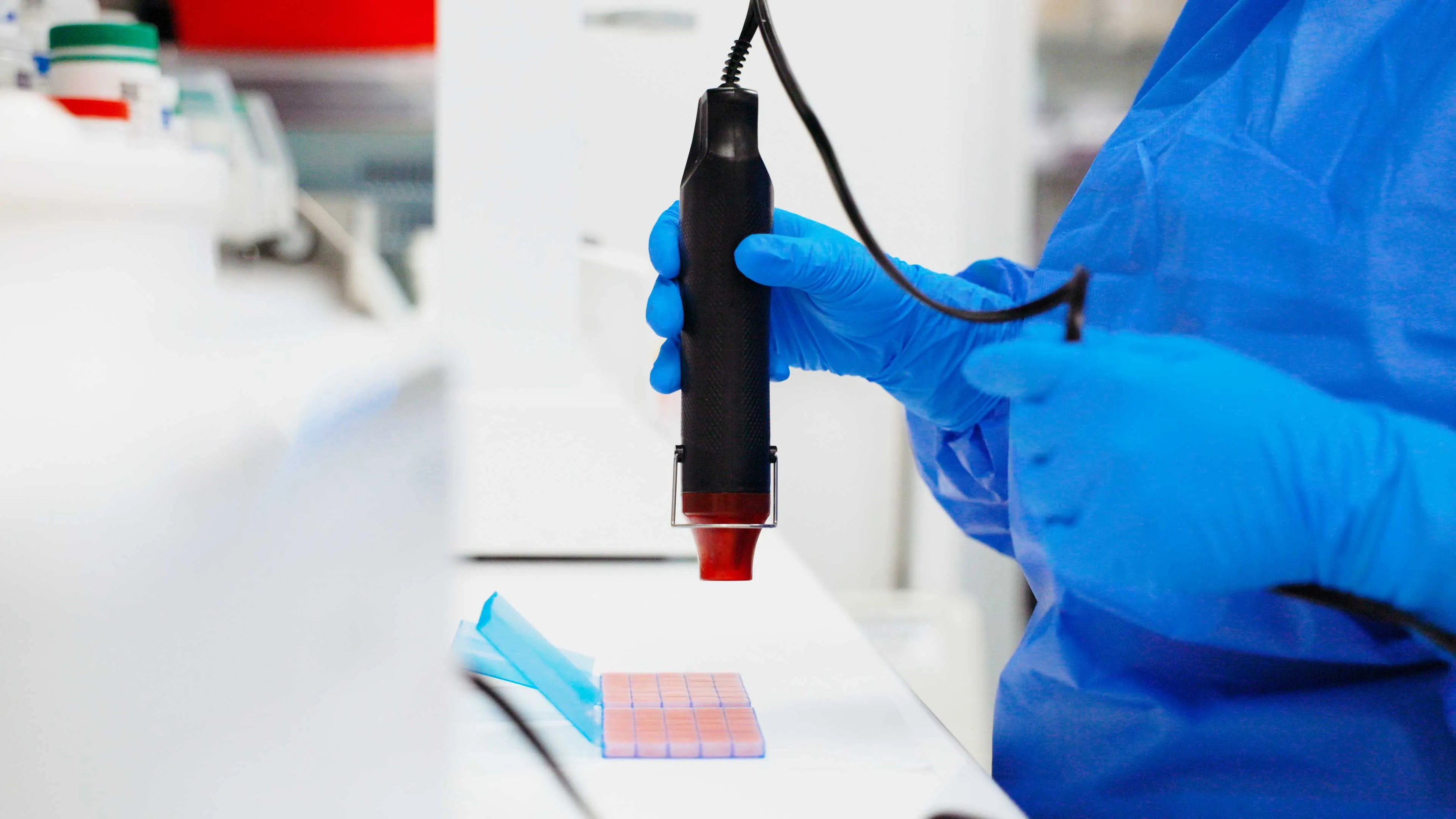 Person in blue gloves using a black handheld device on small lab sample plates in a lab setting.
