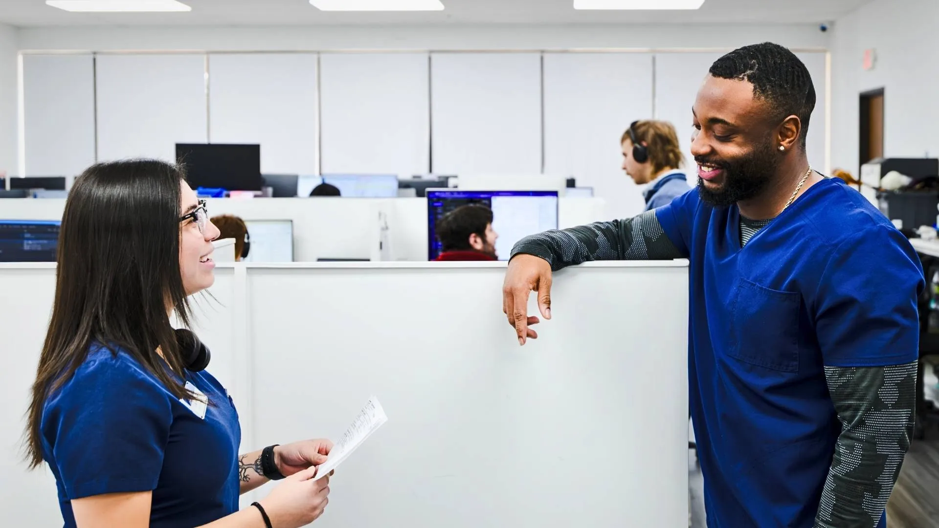 Two healthcare workers in blue scrubs talking and smiling in an office with cubicles and computer screens.