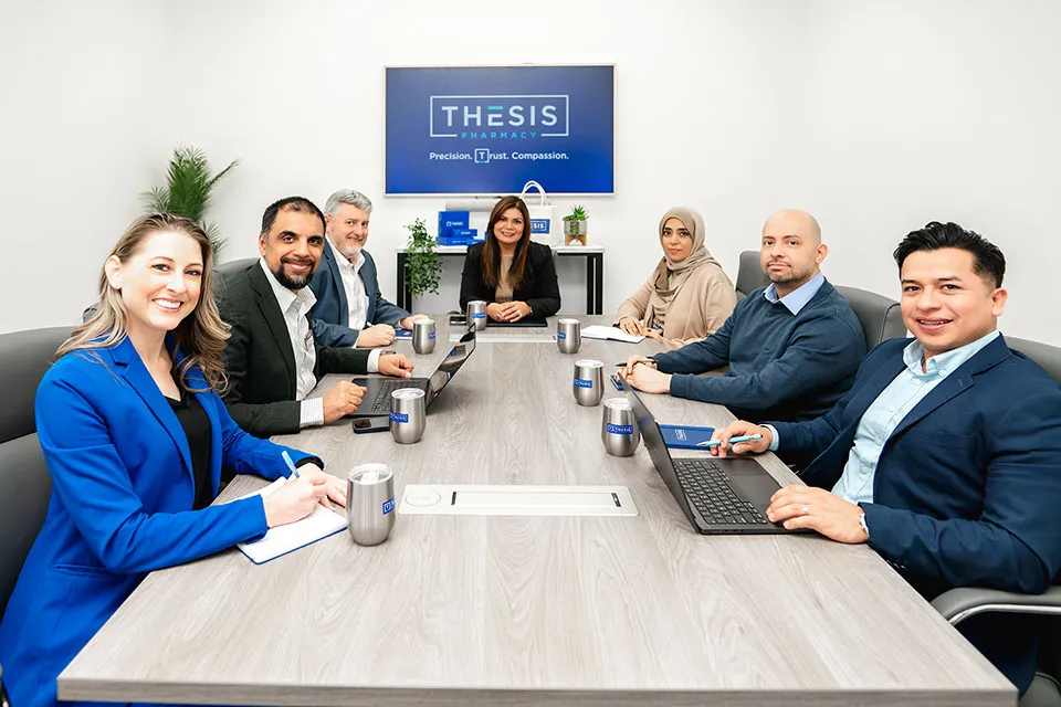 Group of seven diverse professionals in a meeting room with laptops and Thesis Pharmacy sign behind them.