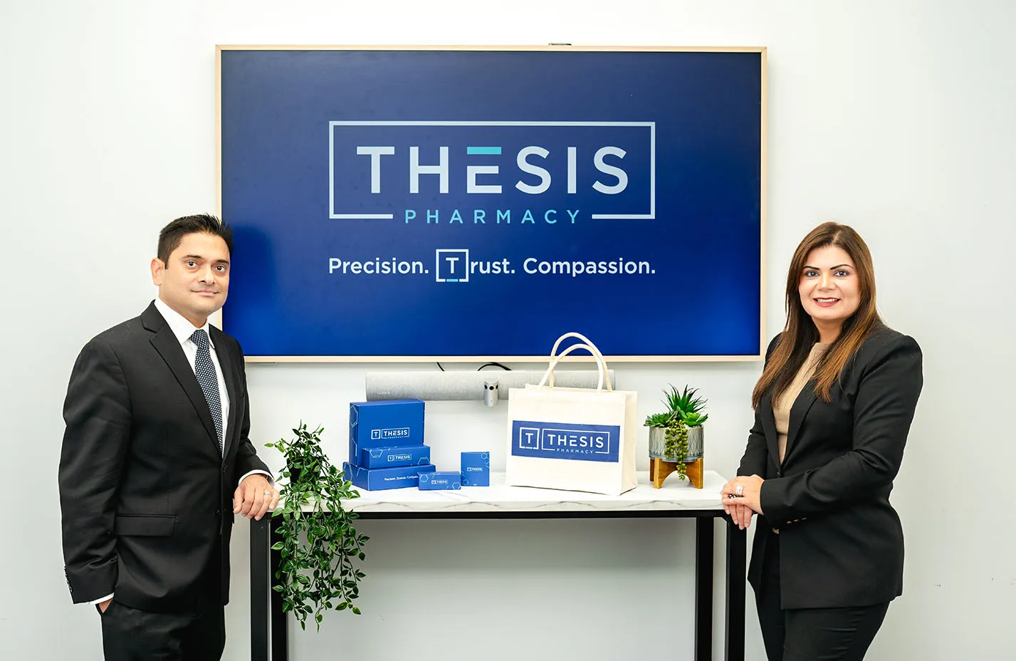 Man and woman in business attire standing beside table with Thesis Pharmacy branded items and a screen.