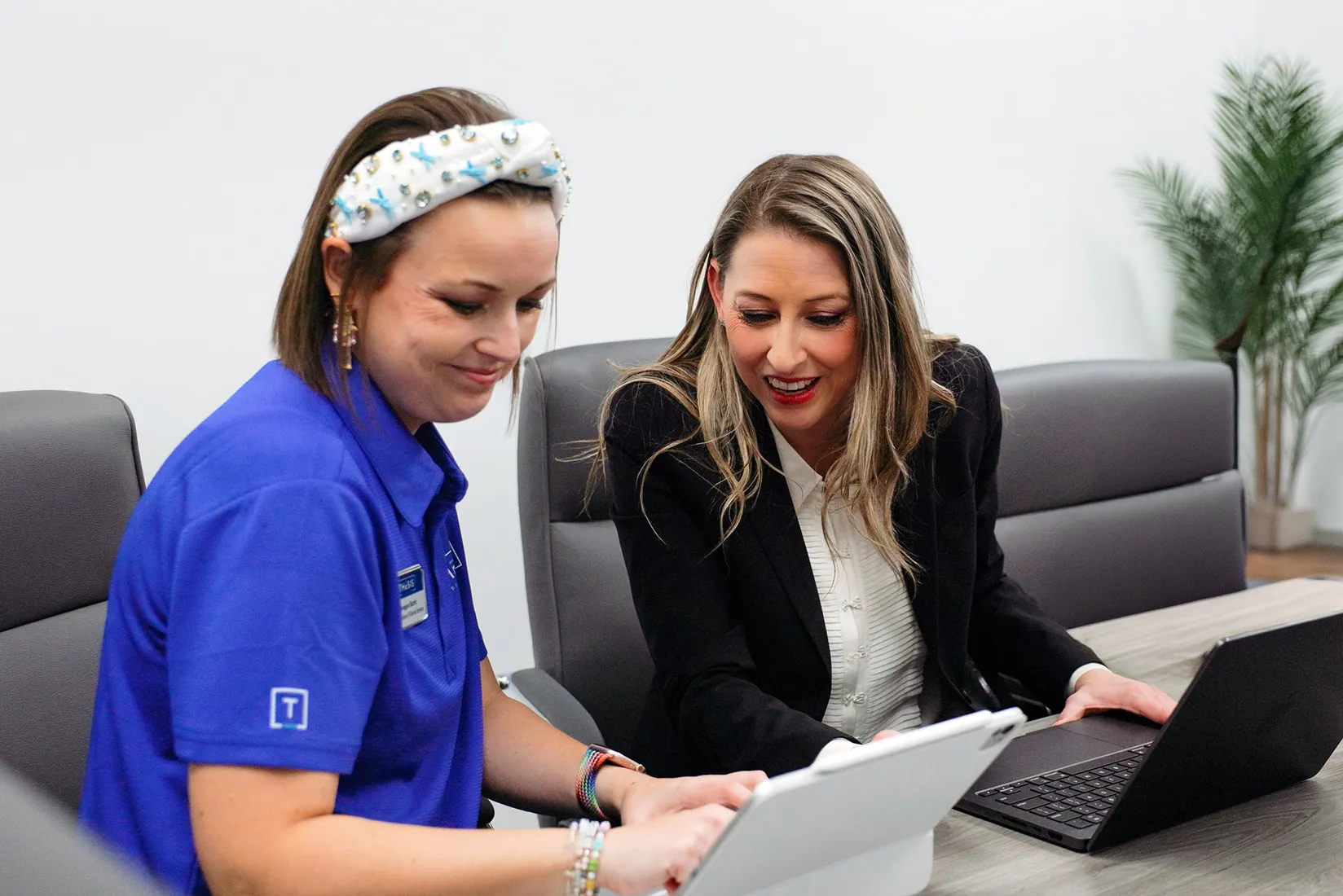 Two women sitting at a table working on a tablet and laptop, smiling and discussing together.
