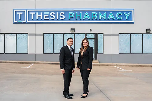 Two professionally dressed people standing outside Thesis Pharmacy building with a blue sign.