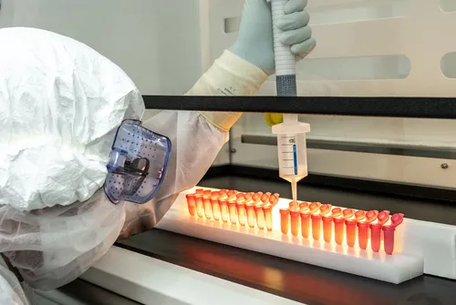 Scientist in protective gear uses pipette to fill rows of glowing test tubes in lab.