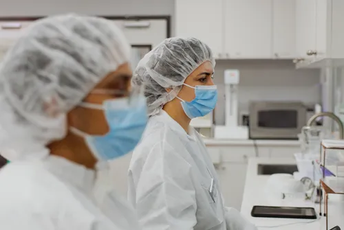 Two healthcare workers wearing hairnets, masks, and protective gowns in a clinical lab setting.