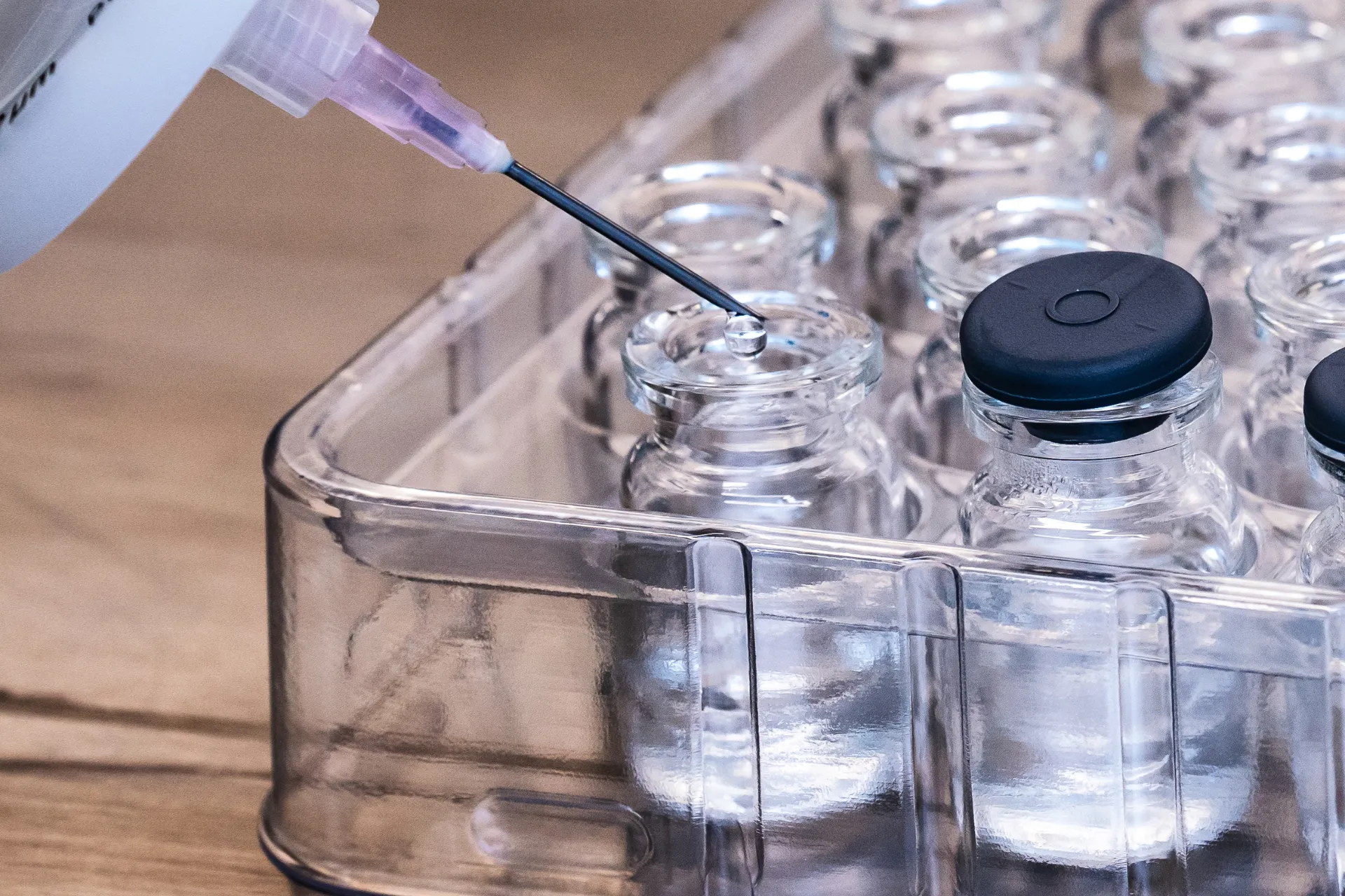 Syringe needle drawing liquid from a clear glass vial among multiple similar vials in a plastic tray.