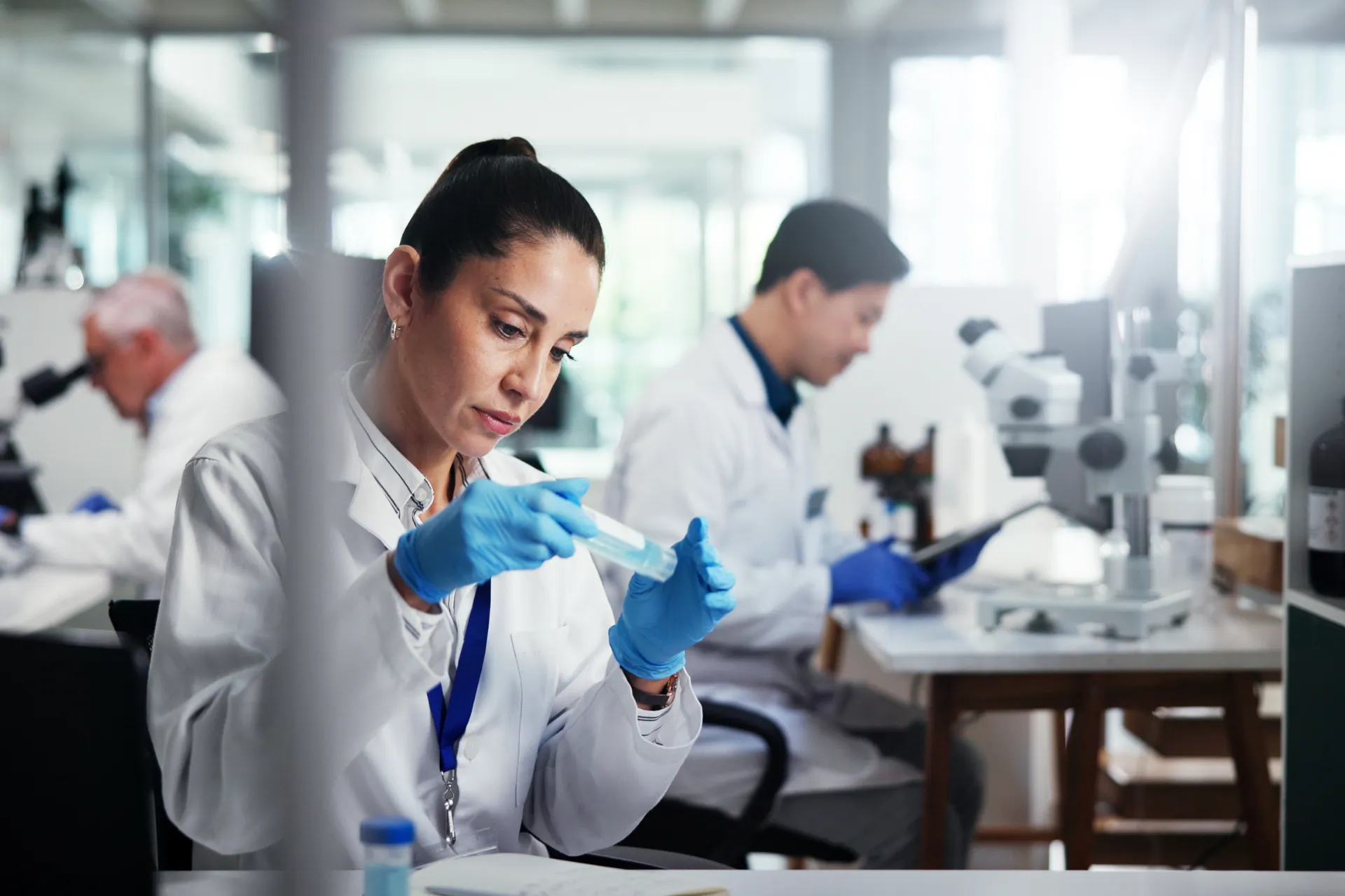 Female scientist wearing gloves examines sample in test tube in a busy modern lab with colleagues.