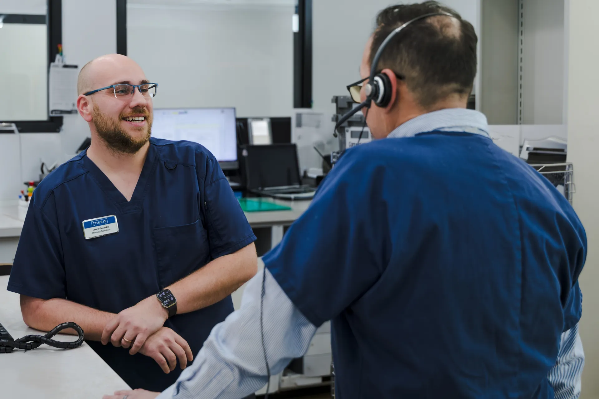 Two pharmacy technicians in navy scrubs chatting at a pharmacy work counter.