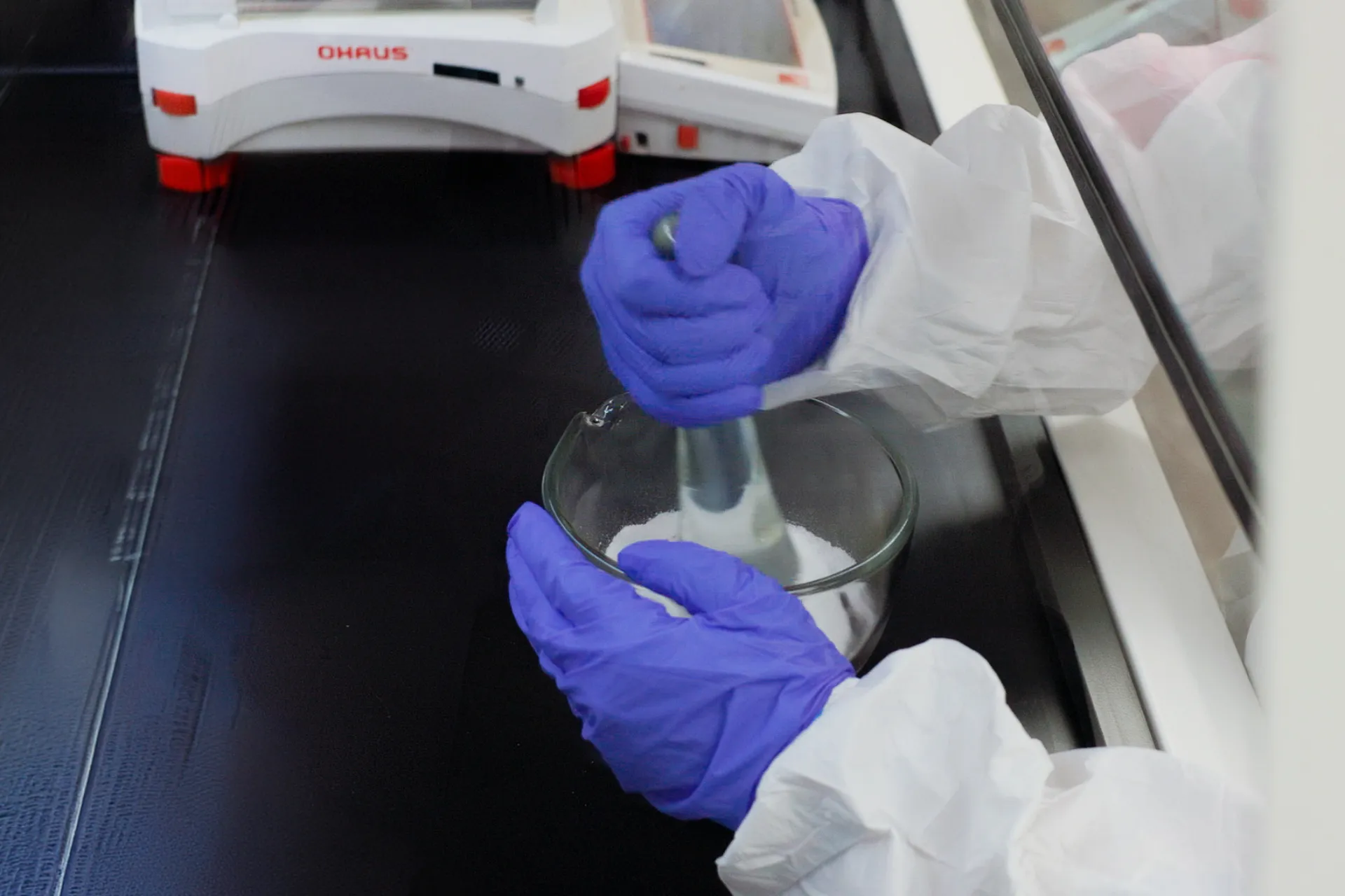 Person wearing blue gloves and white lab coat mixing white powder in glass bowl with mortar and pestle.