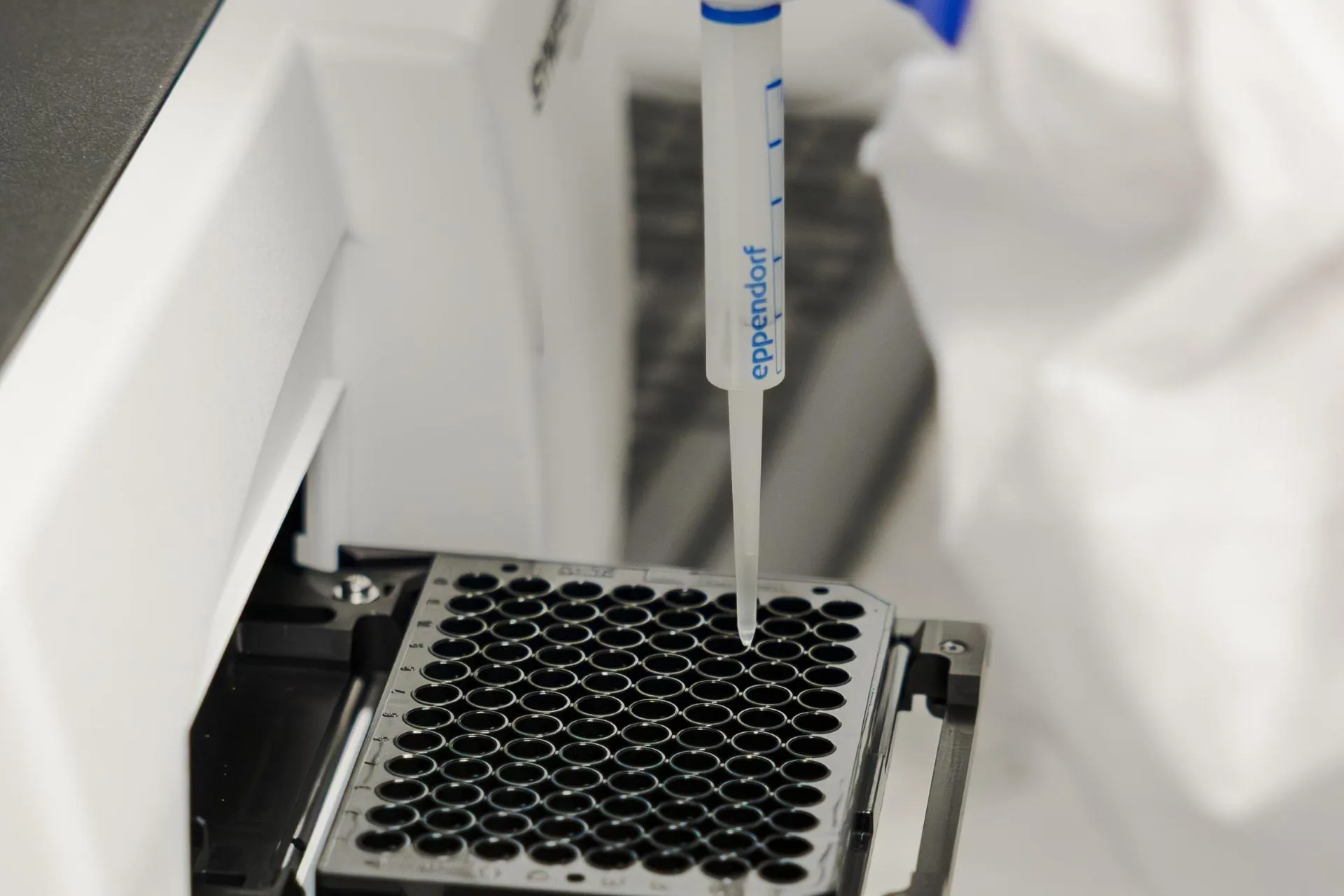 Gloved hand using Eppendorf pipette to add liquid to a well plate in a lab instrument.