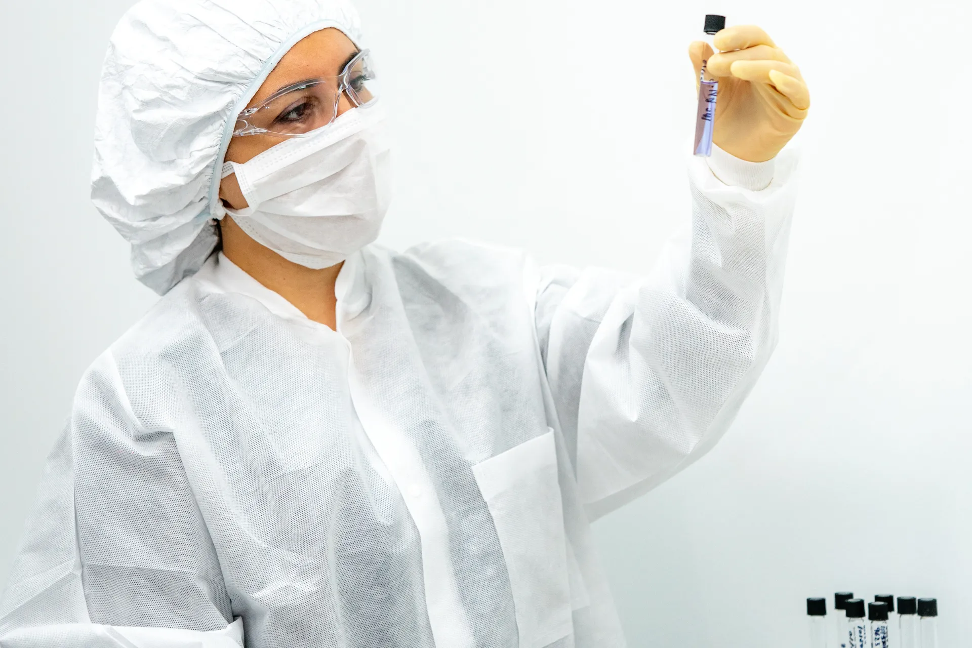 Scientist in protective gear examining a test tube with liquid in a lab setting.