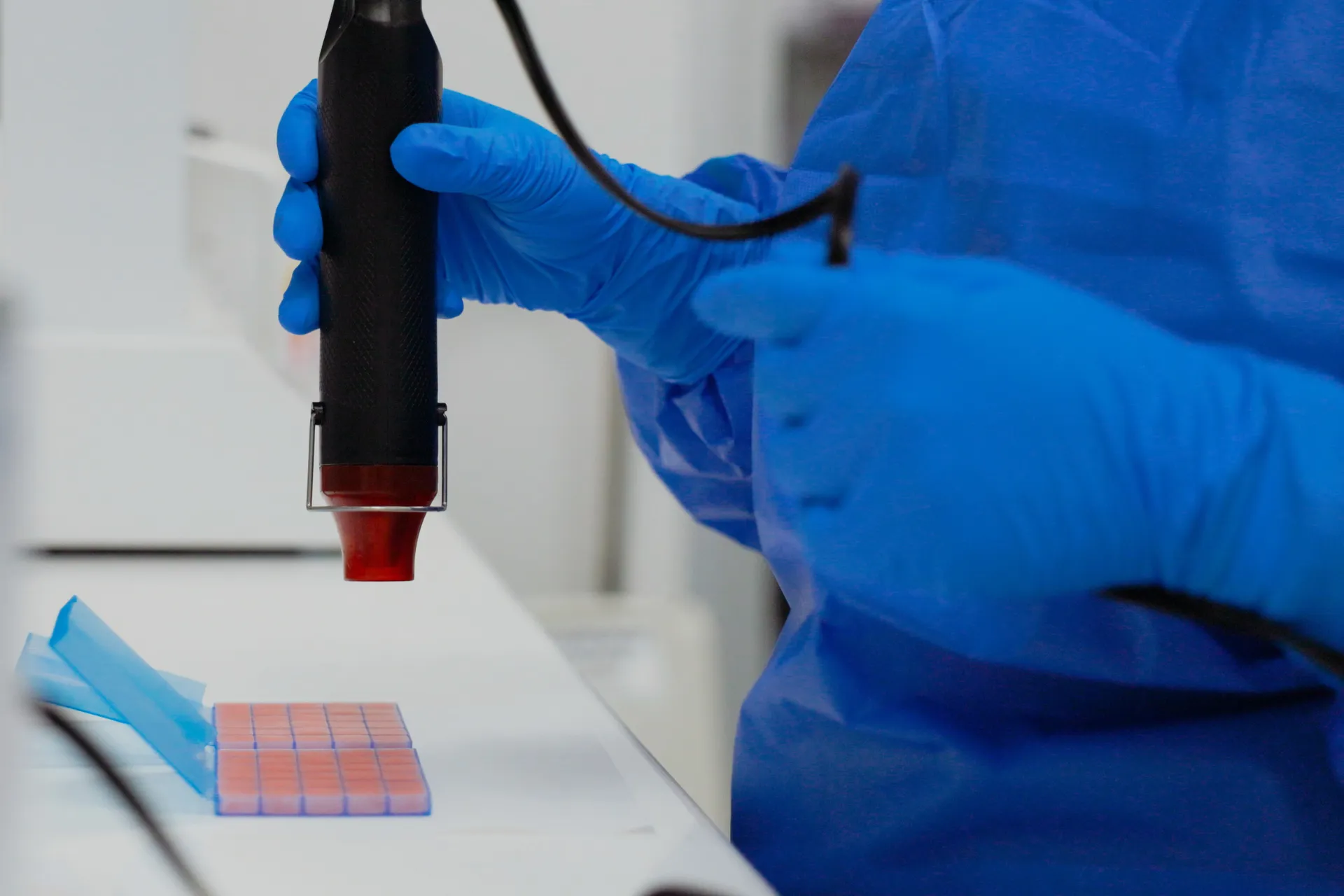 Technician in blue gloves and gown uses a handheld device over pink samples in a lab setting.