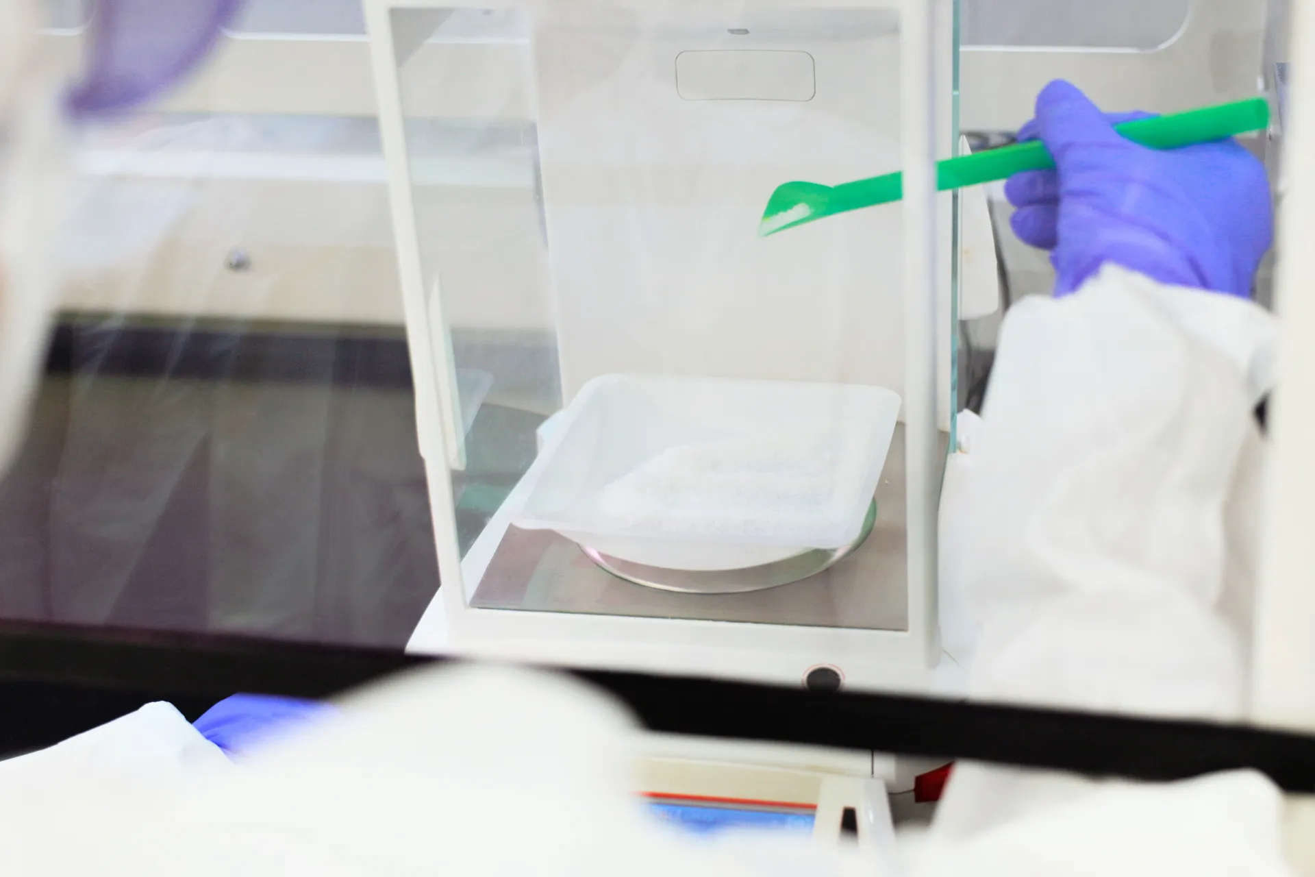 Lab technician in gloves handling white powder on a scale with a green spatula.