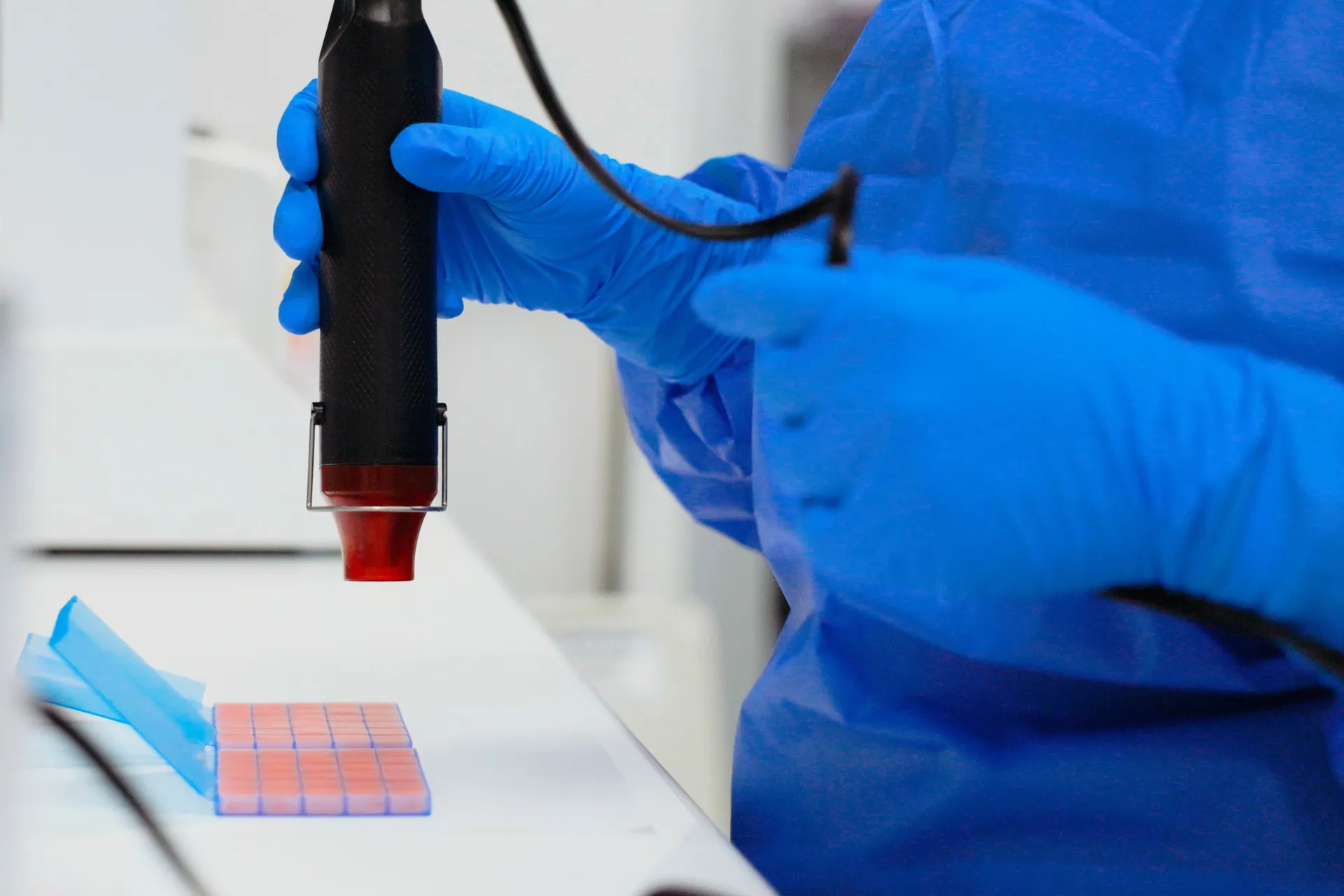 Person in blue gloves and gown holding black device over a grid of reddish samples in lab.