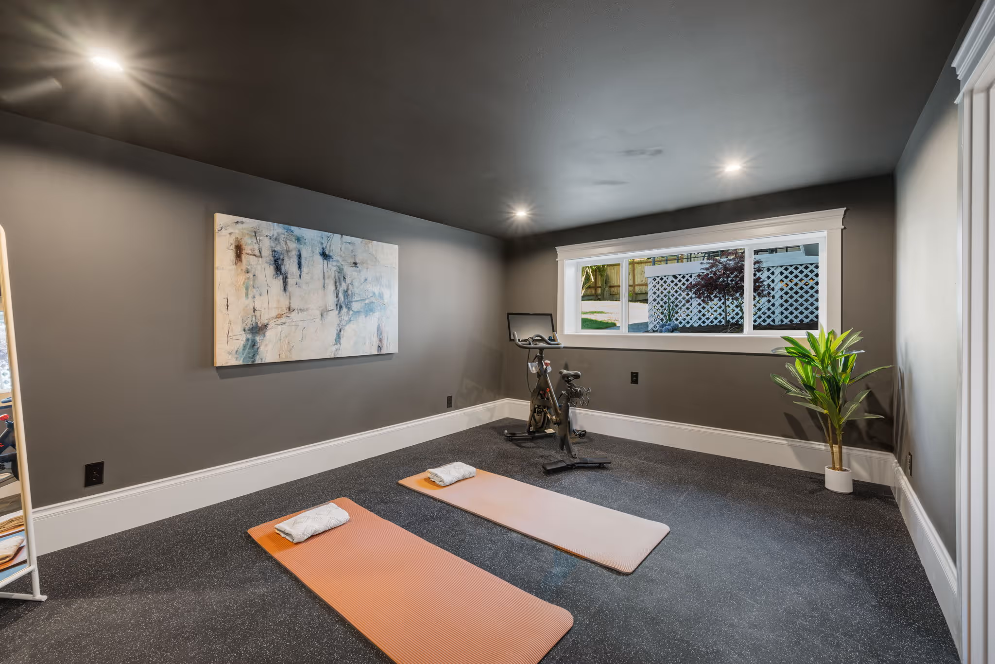 Home workout room with two yoga mats, exercise bike, abstract wall art, and a potted plant by window.