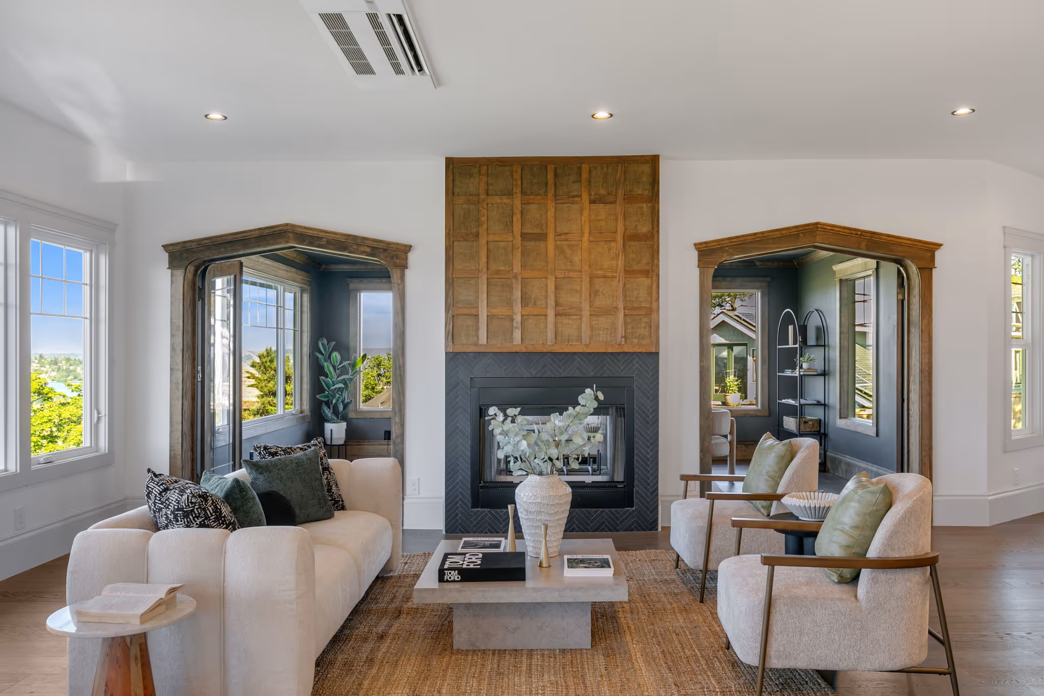 Modern living room with white sofa, two armchairs, a coffee table with vase, and a fireplace between two wood-framed mirrors.