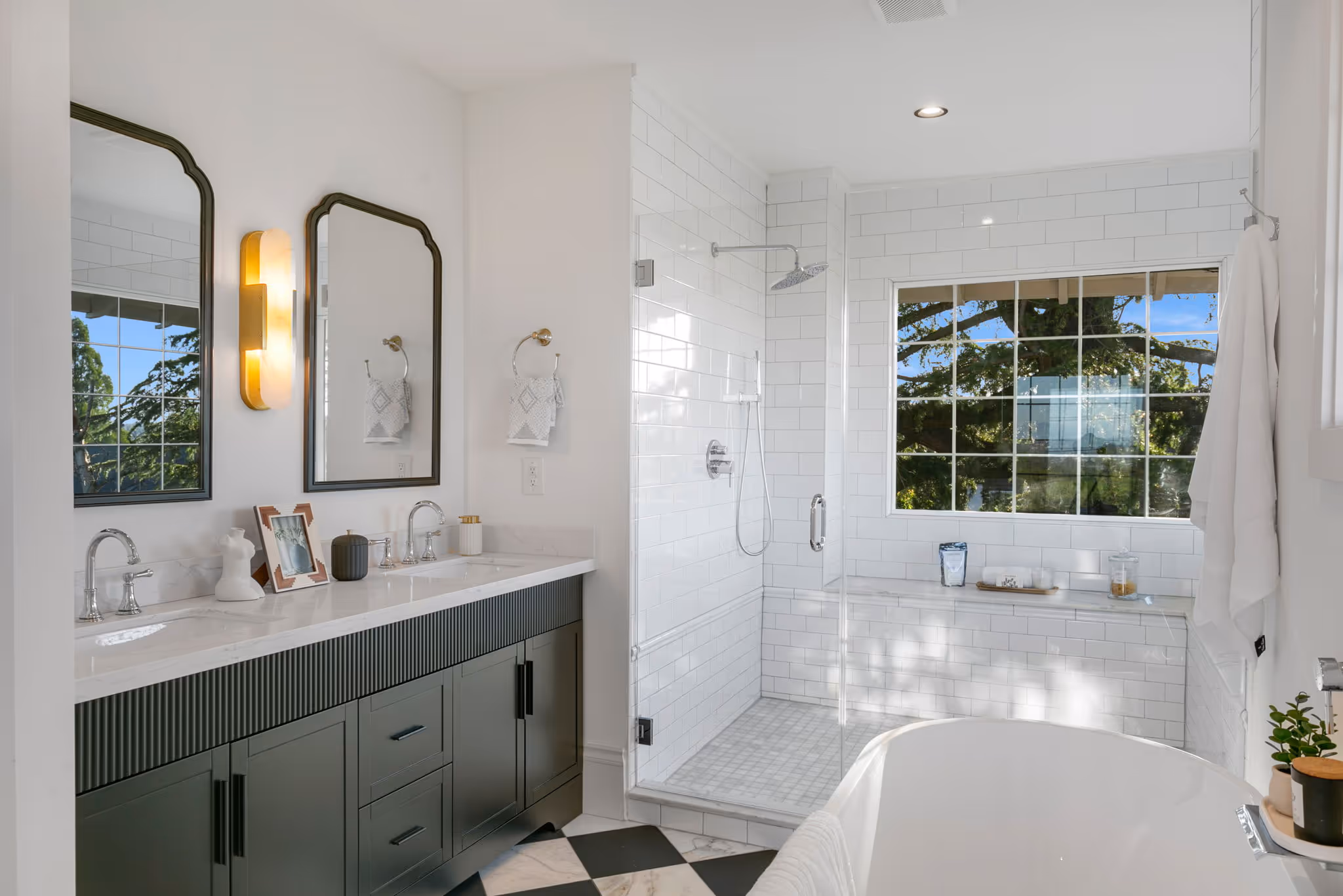 Modern bathroom with dark green double vanity, large mirrors, glass shower, and white freestanding tub.