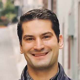 Smiling man with dark hair standing outside on a blurred street background.