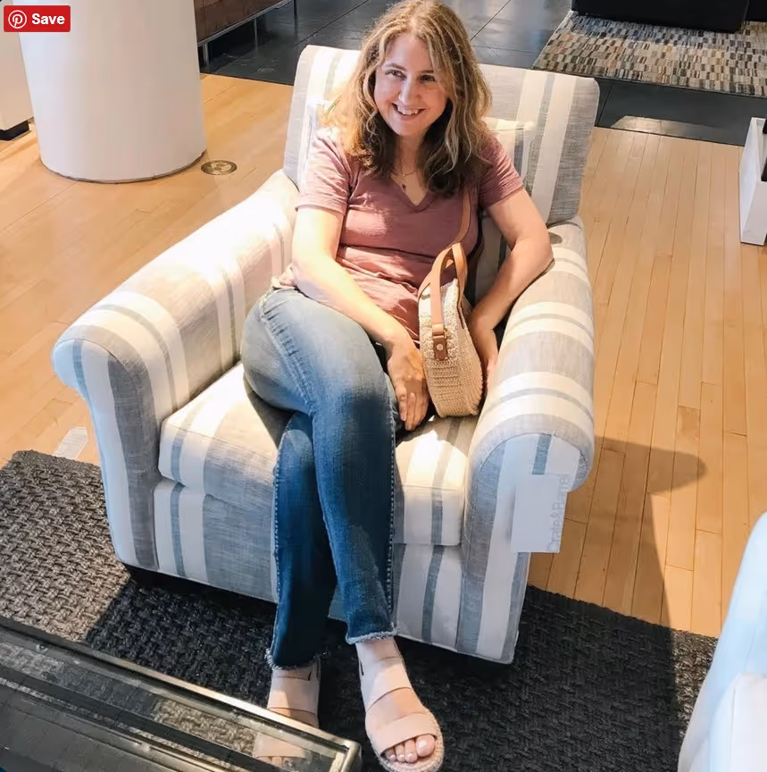 Smiling woman in pink shirt and jeans sitting cross-legged on striped armchair holding a woven bag.