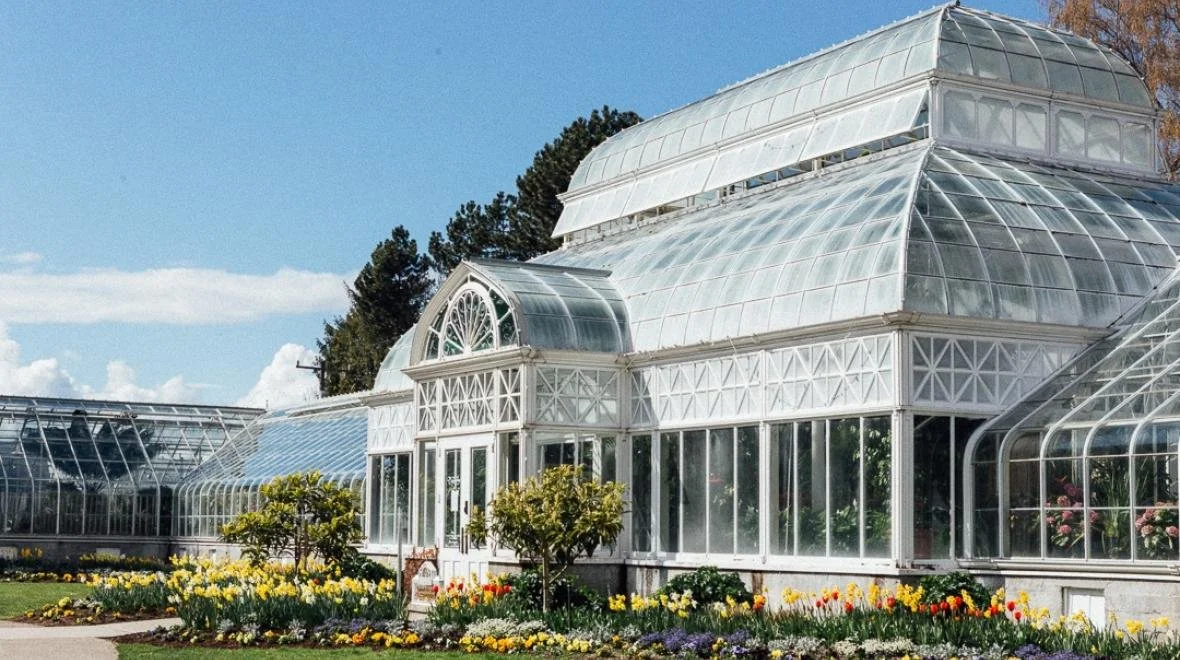 Glass greenhouse building with arched windows surrounded by colorful flower beds and small trees.