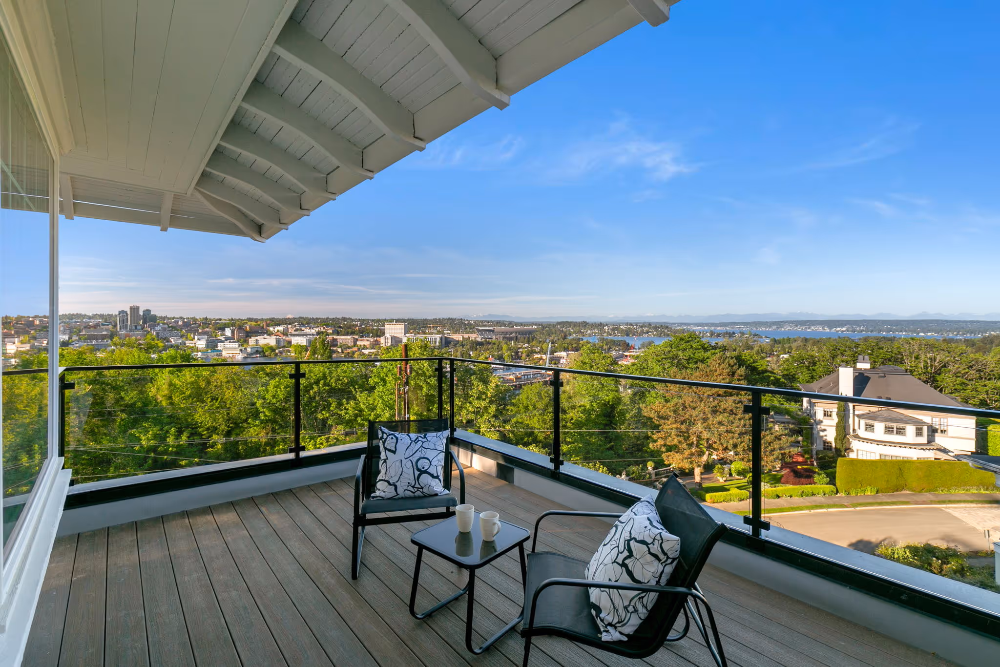 Balcony with two black chairs, floral cushions, small table with cups, overlooking cityscape under clear blue sky.